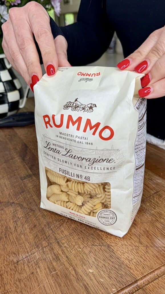 A person with red nail polish holds a white bag of Rummo Fusilli No. 48 pasta on a wooden surface. Perfect for crafting an Easy Italian Pasta Salad, the bag features a clear window displaying the spiral-shaped pasta inside.