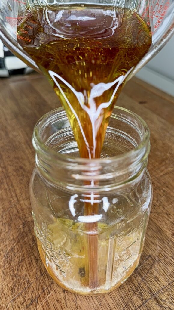 A close-up of amber-colored liquid, perfect for dressing an Easy Italian Pasta Salad, being poured from a glass measuring cup into a clear mason jar on a wooden surface.