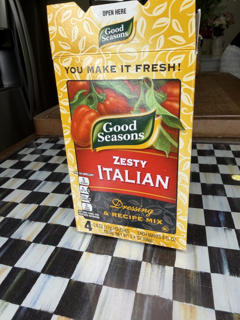 A box of Good Seasons Zesty Italian Dressing & Recipe Mix sits on a black-and-white checkered table. The yellow box, perfect for Easy Italian Pasta Salad, features images of tomatoes and green leaves with text advertising its freshness.