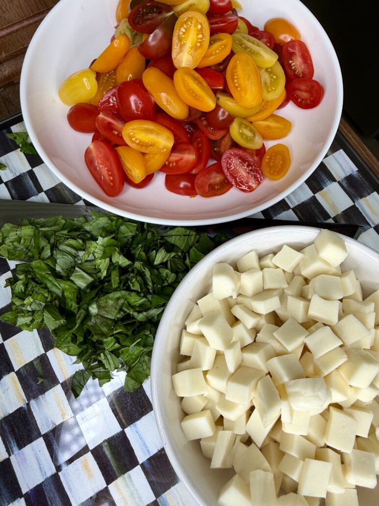 A bowl of halved yellow and red cherry tomatoes, cubed mozzarella cheese, and a pile of chopped fresh basil on a checkered cloth—perfect ingredients for an Easy Italian Pasta Salad.