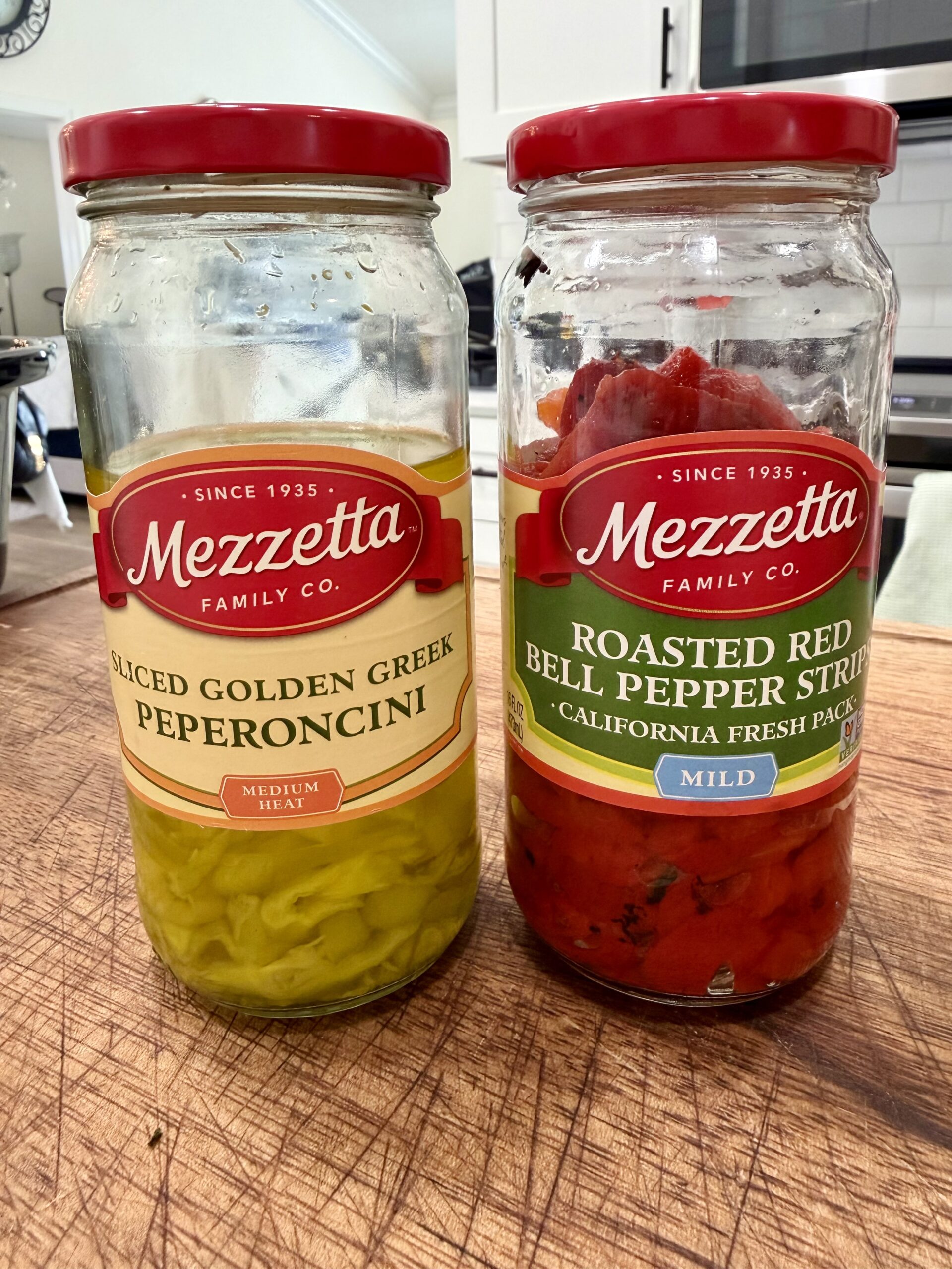 Two open jars of Mezzetta brand peppers on a wooden surface: one contains sliced golden Greek pepperoncini, and the other contains roasted red bell pepper strips. A kitchen counter and stove are in the background.