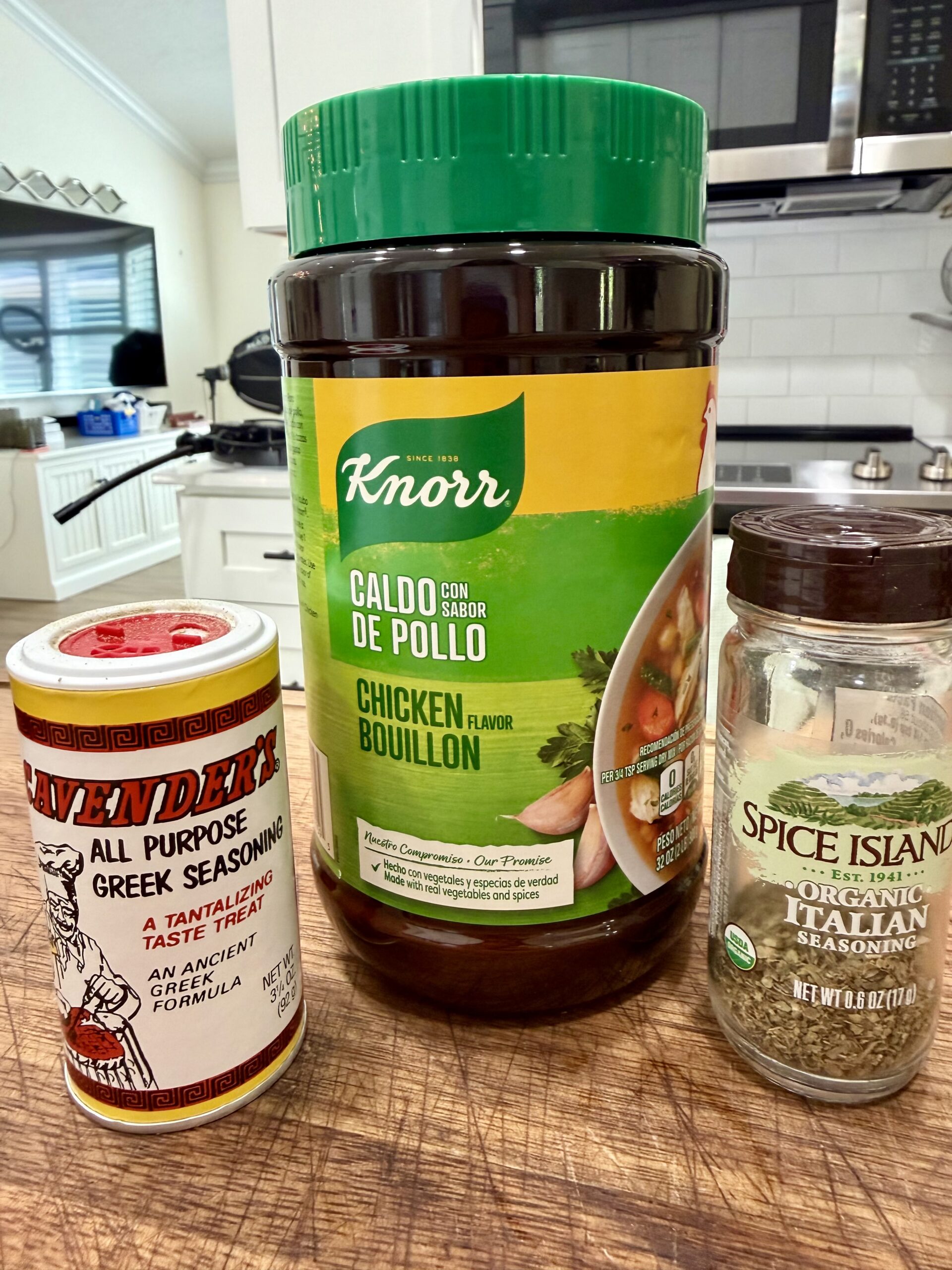 Three spice containers—Lawrys Greek Seasoning, Knorr Chicken Bouillon, and Spice Islands Organic Italian Seasoning—stand on a wooden countertop in a kitchen with white cabinets and a stove in the background.