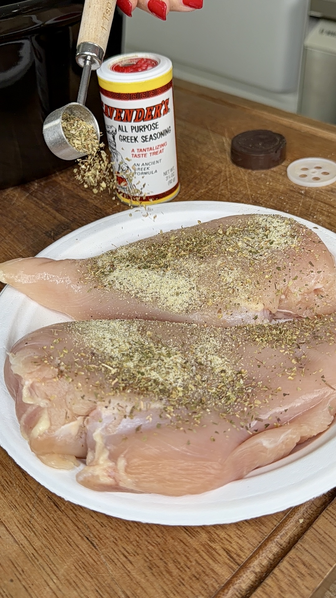 A hand sprinkles dried seasoning onto two raw chicken breasts on a white plate. A container of Greek seasoning sits nearby on a wooden surface.