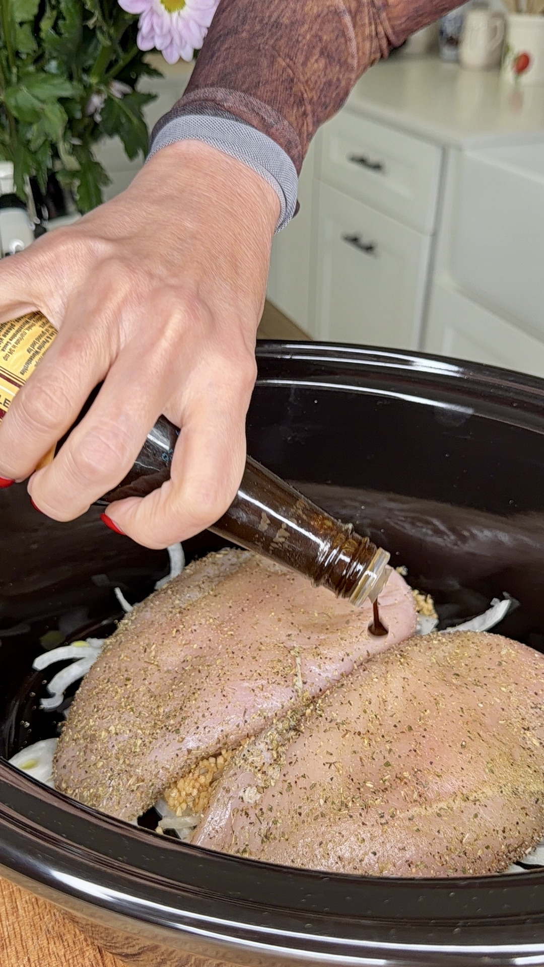 A hand pours a dark liquid from a bottle onto seasoned raw turkey breasts placed over sliced onions in a black slow cooker, with a kitchen counter and cabinets in the background.