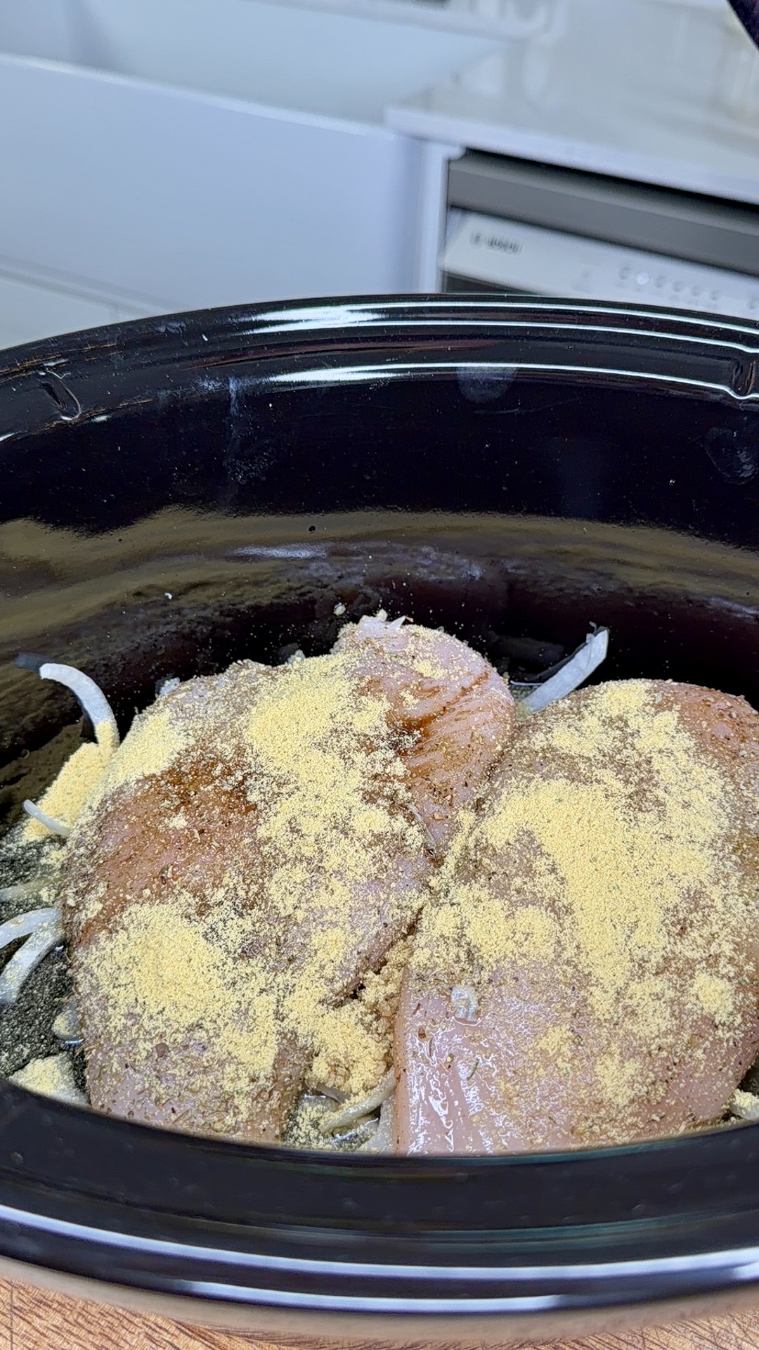 Raw chicken breasts covered in seasoning and placed on top of sliced onions inside a black slow cooker, ready to be cooked. Kitchen counters and a dishwasher are visible in the background.