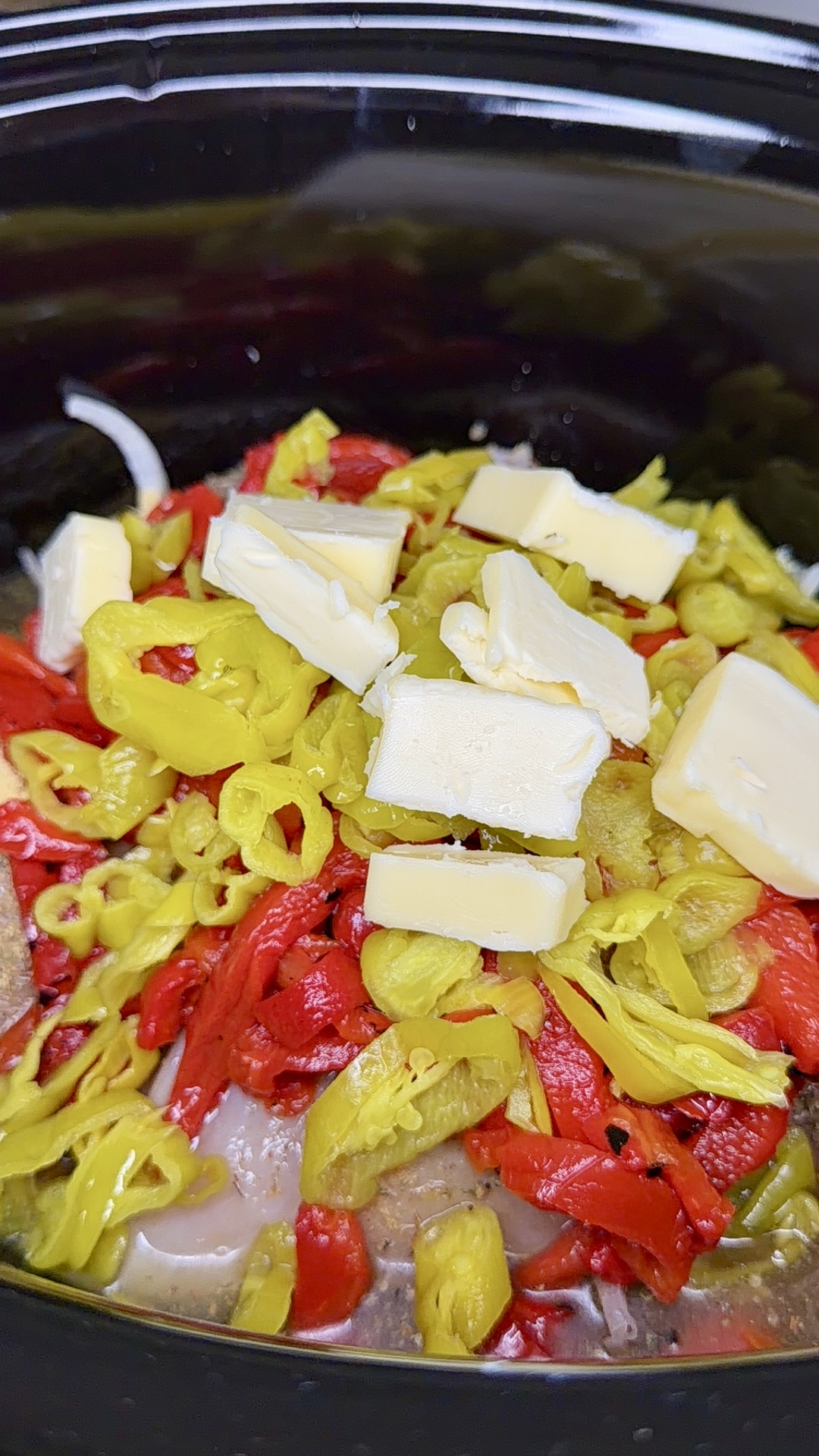 Sliced yellow peppers, red peppers, and several pats of butter are layered on top of meat and onions inside a black slow cooker.