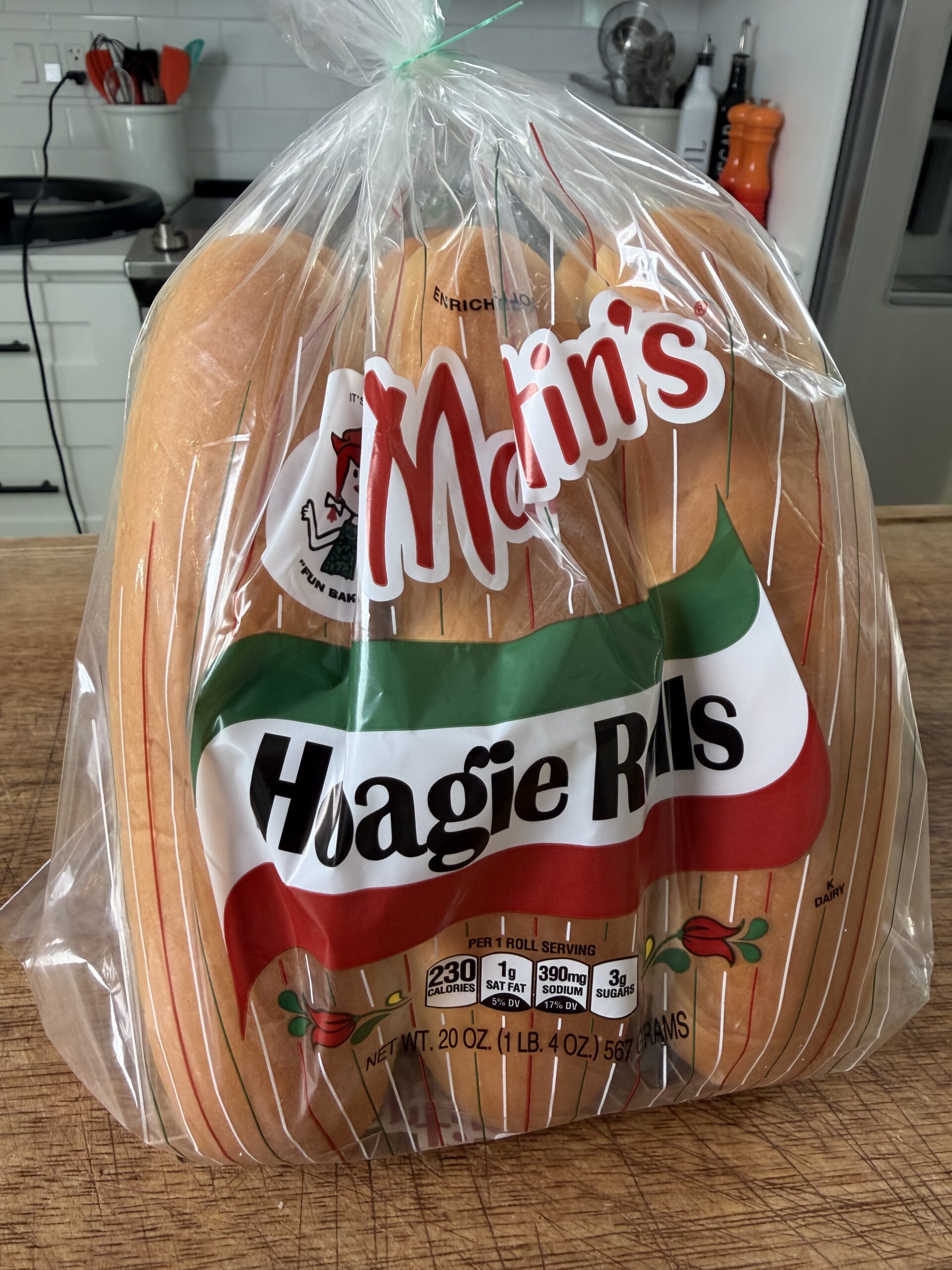 A sealed plastic bag of Martins Hoagie Rolls sits on a wooden countertop in a modern kitchen, ready to be paired with an Easy Crockpot Chicken Philly Cheesesteak. White cabinets, utensils, and appliances fill the background.