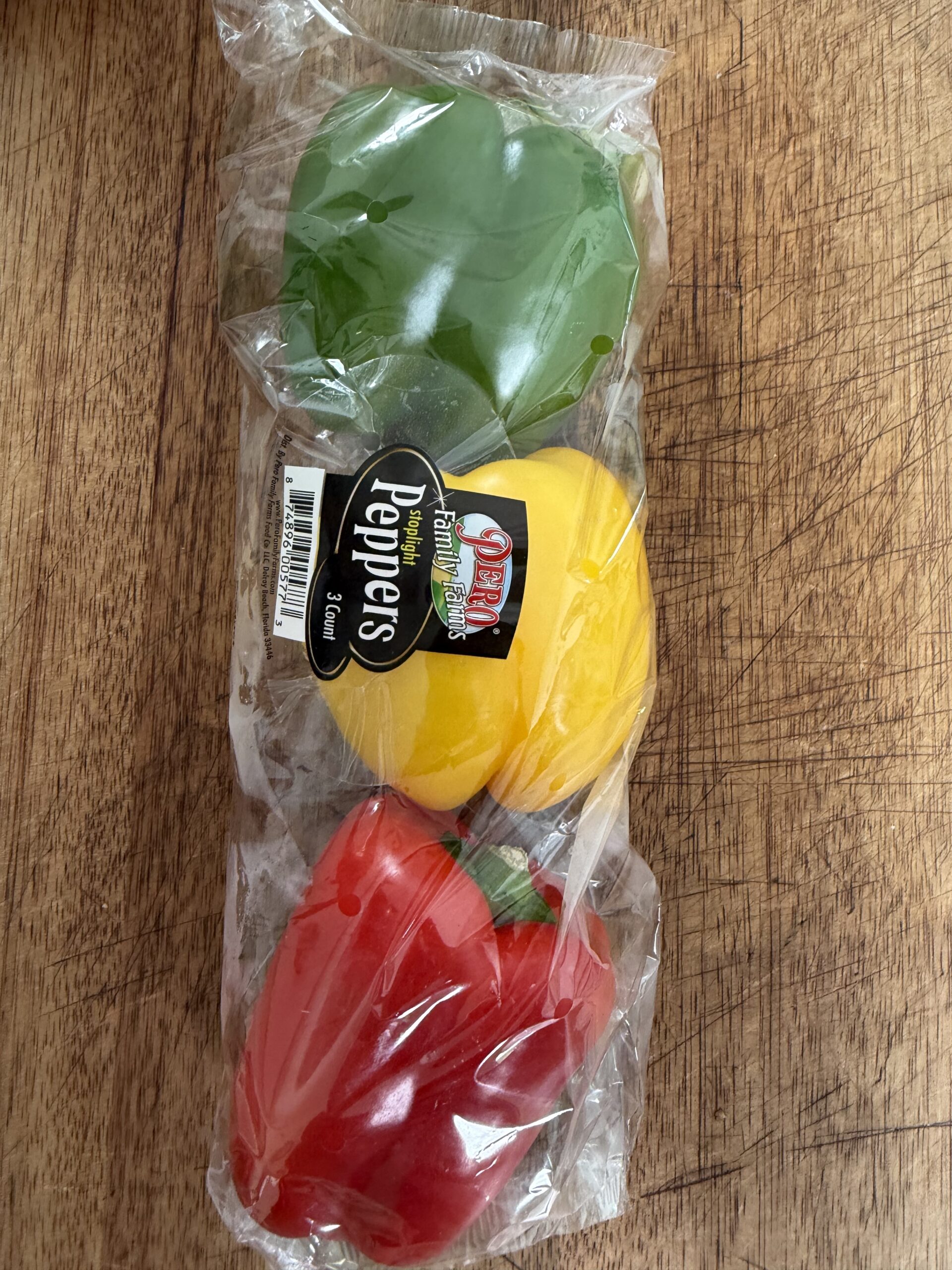 Three bell peppers—green, yellow, and red—are packaged together in clear plastic wrapping on a wooden surface. Perfect for recipes like Easy Crockpot Philly Cheesesteaks, the label reads Peppers with branding and a barcode visible.