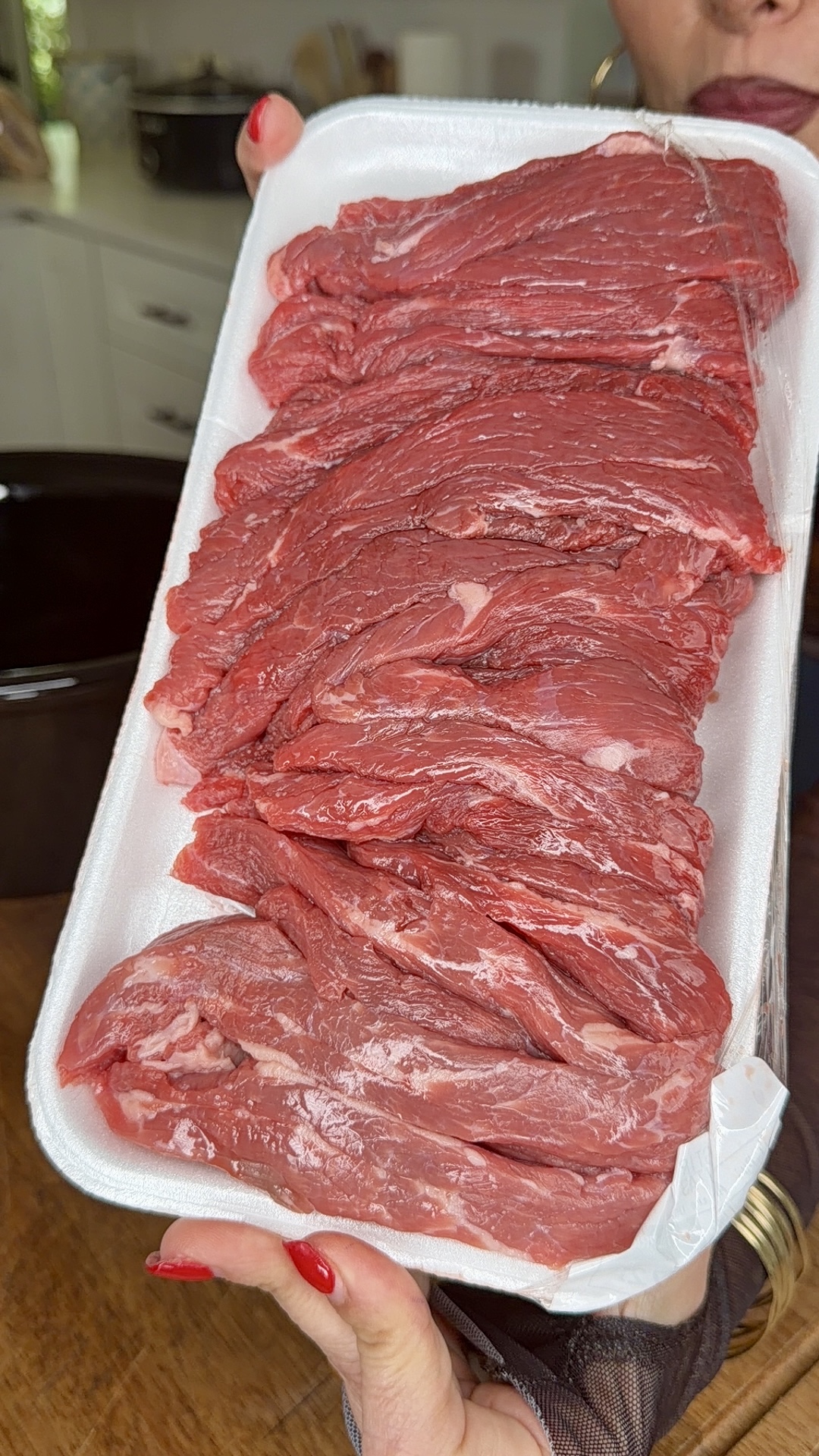 A person with red painted nails holds a styrofoam tray of raw, thinly sliced red beef—perfect for Easy Crockpot Philly Cheesesteaks—partially covered with plastic wrap, in a kitchen setting.
