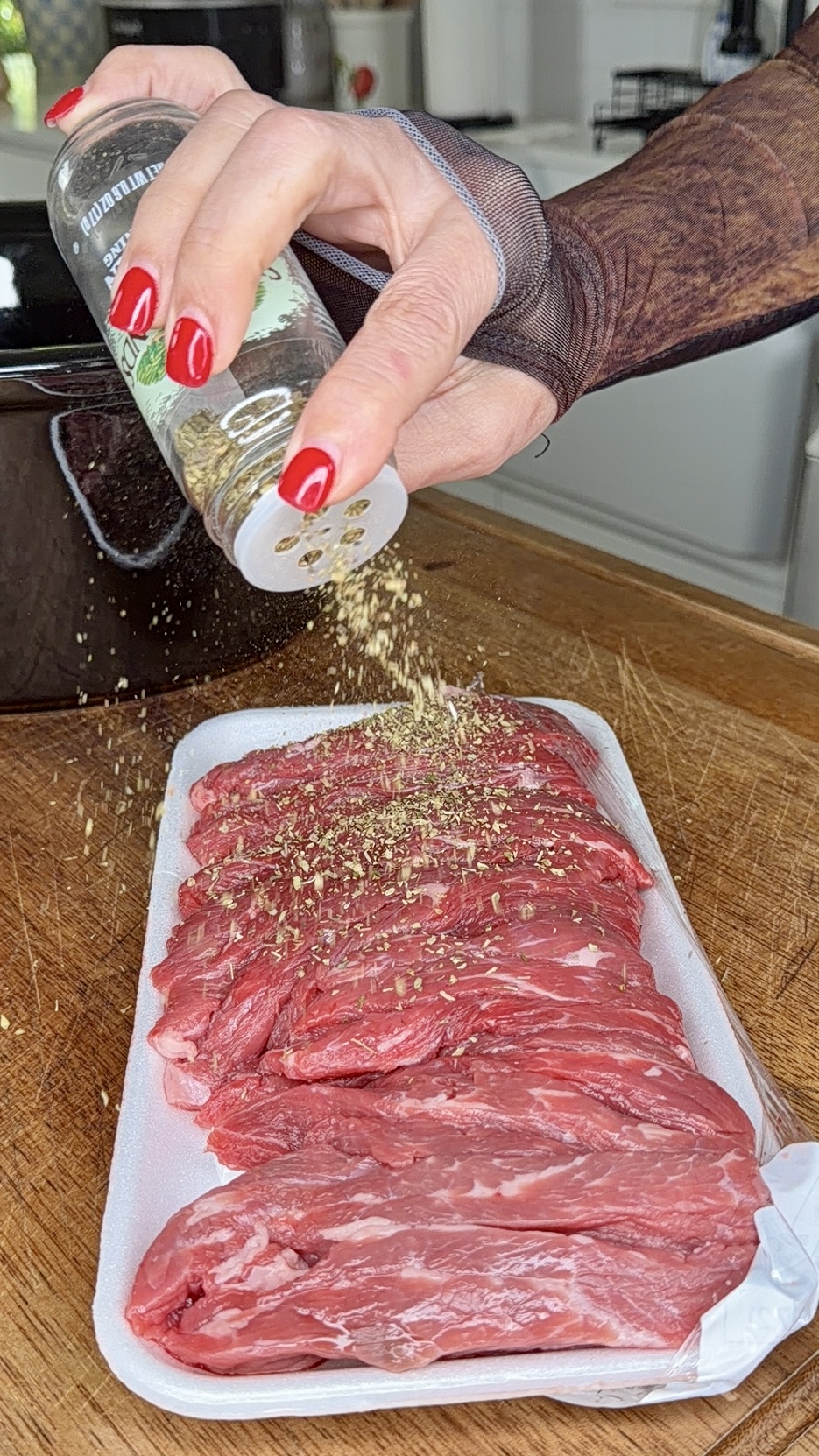 A hand with red nails sprinkles seasoning from a bottle onto raw strips of beef—perfect for Easy Crockpot Philly Cheesesteaks—in a white foam tray on a wooden cutting board in a kitchen.