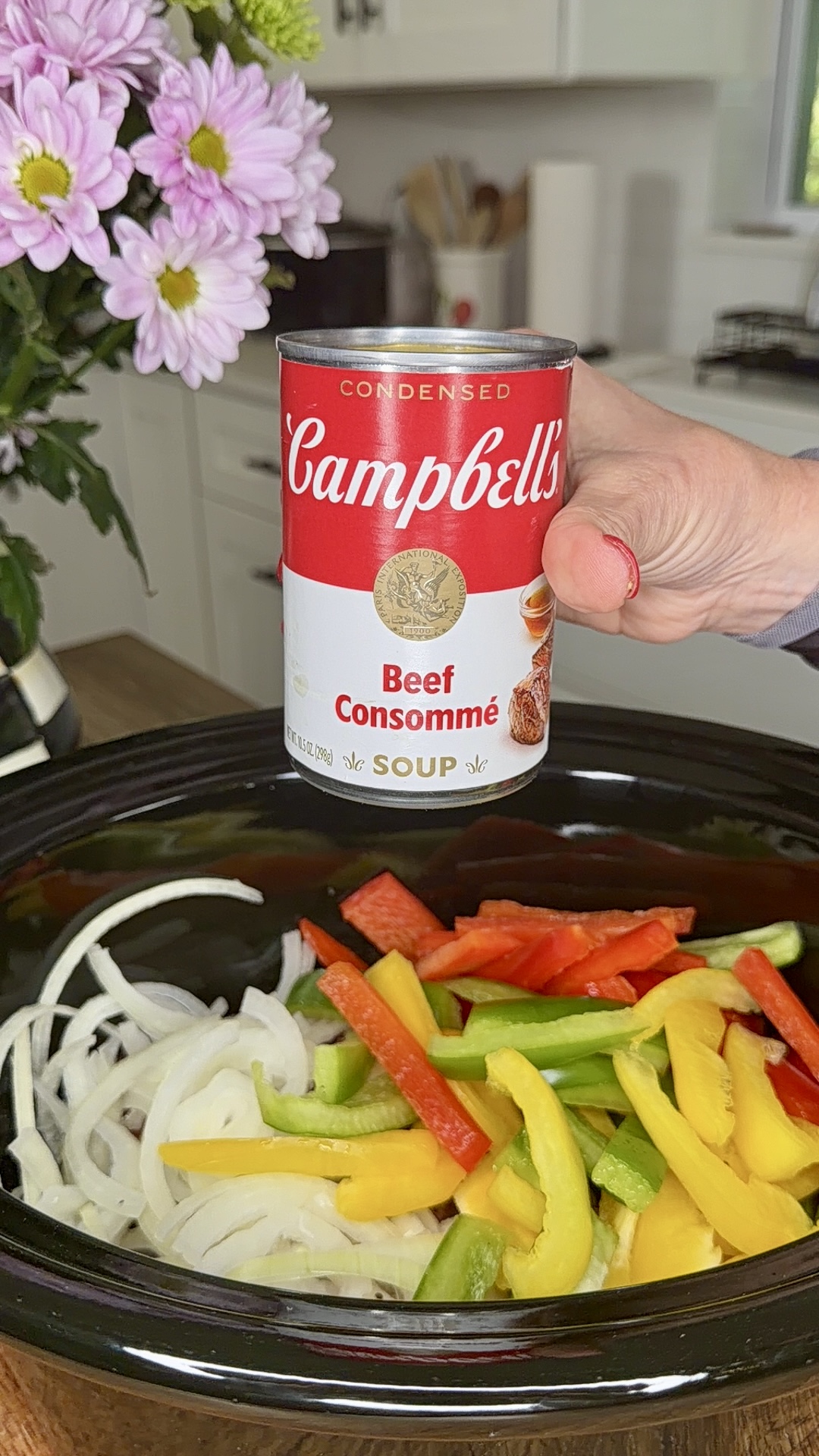A hand holds a can of Campbell’s Beef Consommé soup over a slow cooker filled with sliced onions and colorful bell peppers—perfect for making Easy Crockpot Philly Cheesesteaks. Flowers and a kitchen counter are visible in the background.