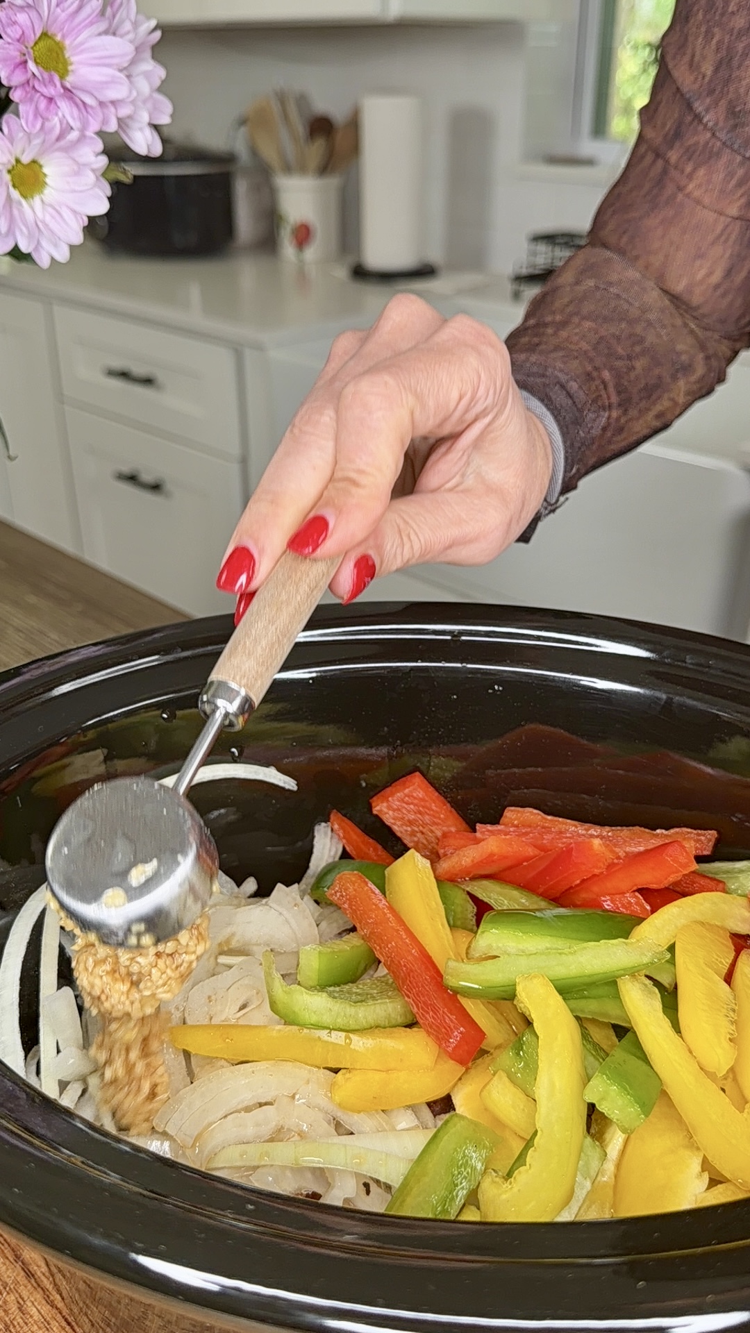 A hand with red nails adds minced garlic to a slow cooker filled with sliced onions and colorful bell peppers—prepping Easy Crockpot Philly Cheesesteaks in a bright kitchen.