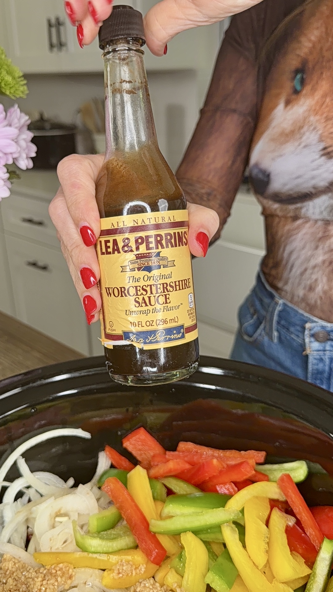 A person with red-painted nails holds a bottle of Lea & Perrins Worcestershire Sauce over a slow cooker filled with sliced onions and colorful bell peppers—perfect for making Easy Crockpot Philly Cheesesteaks in the kitchen.
