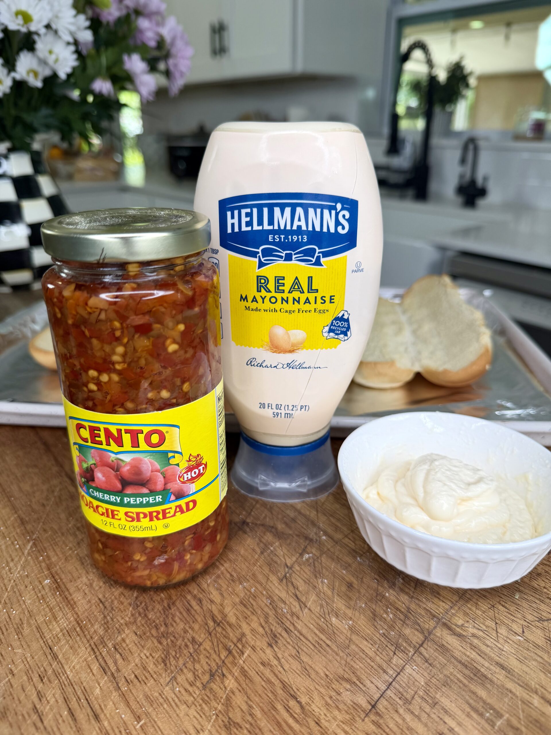 A jar of Cento hot cherry pepper spread, Hellmann’s Real Mayonnaise, and sliced bread are displayed on a wooden kitchen counter—perfect ingredients to enjoy with an Easy Crockpot Chicken Philly Cheesesteak. A kitchen sink and flowers are in the background.