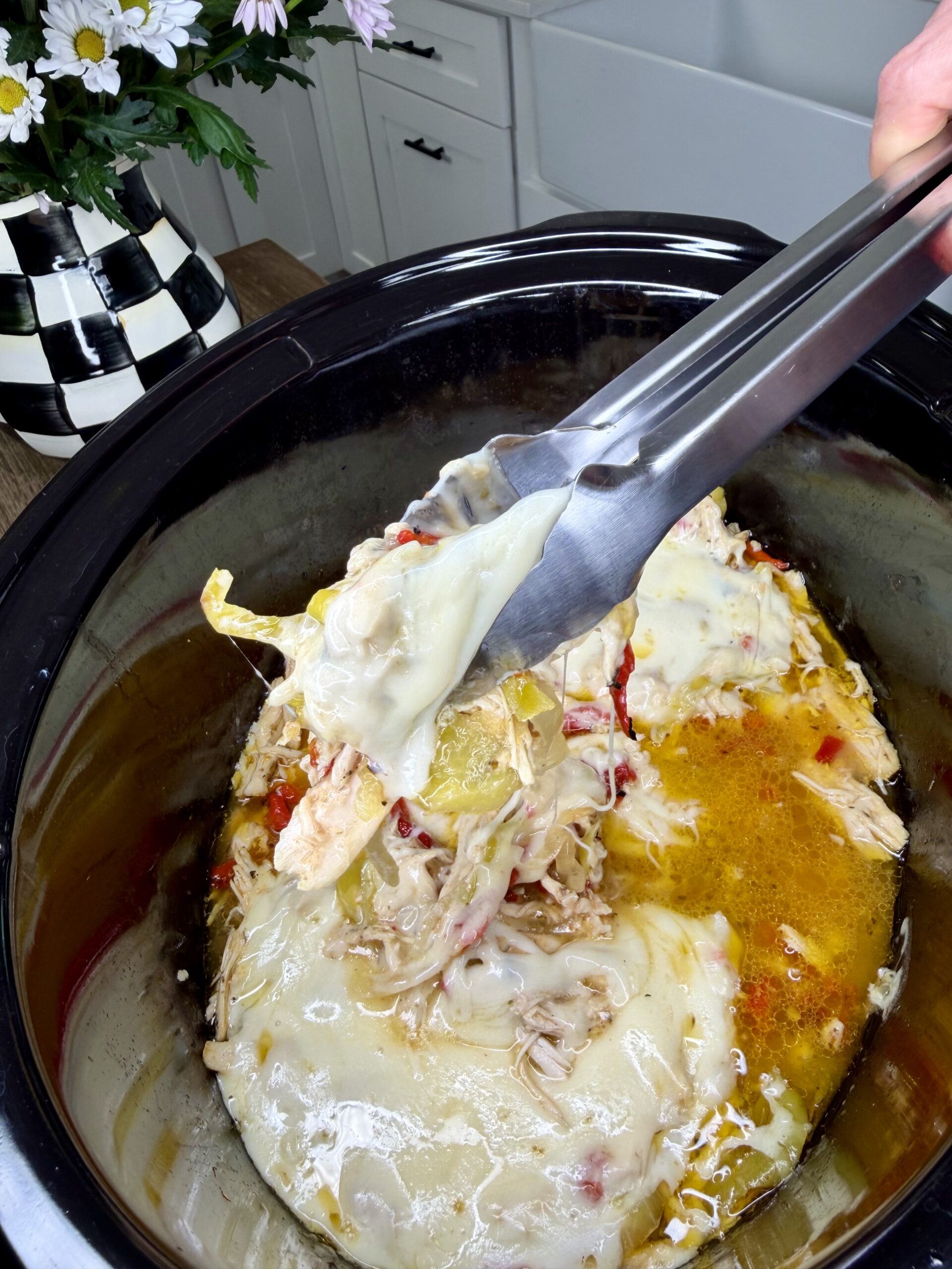 A close-up of an Easy Crockpot Chicken Philly Cheesesteak shows melted cheese, shredded chicken, and veggies in a slow cooker. A hand lifts a portion with tongs while a black-and-white checkered vase with flowers decorates the counter nearby.