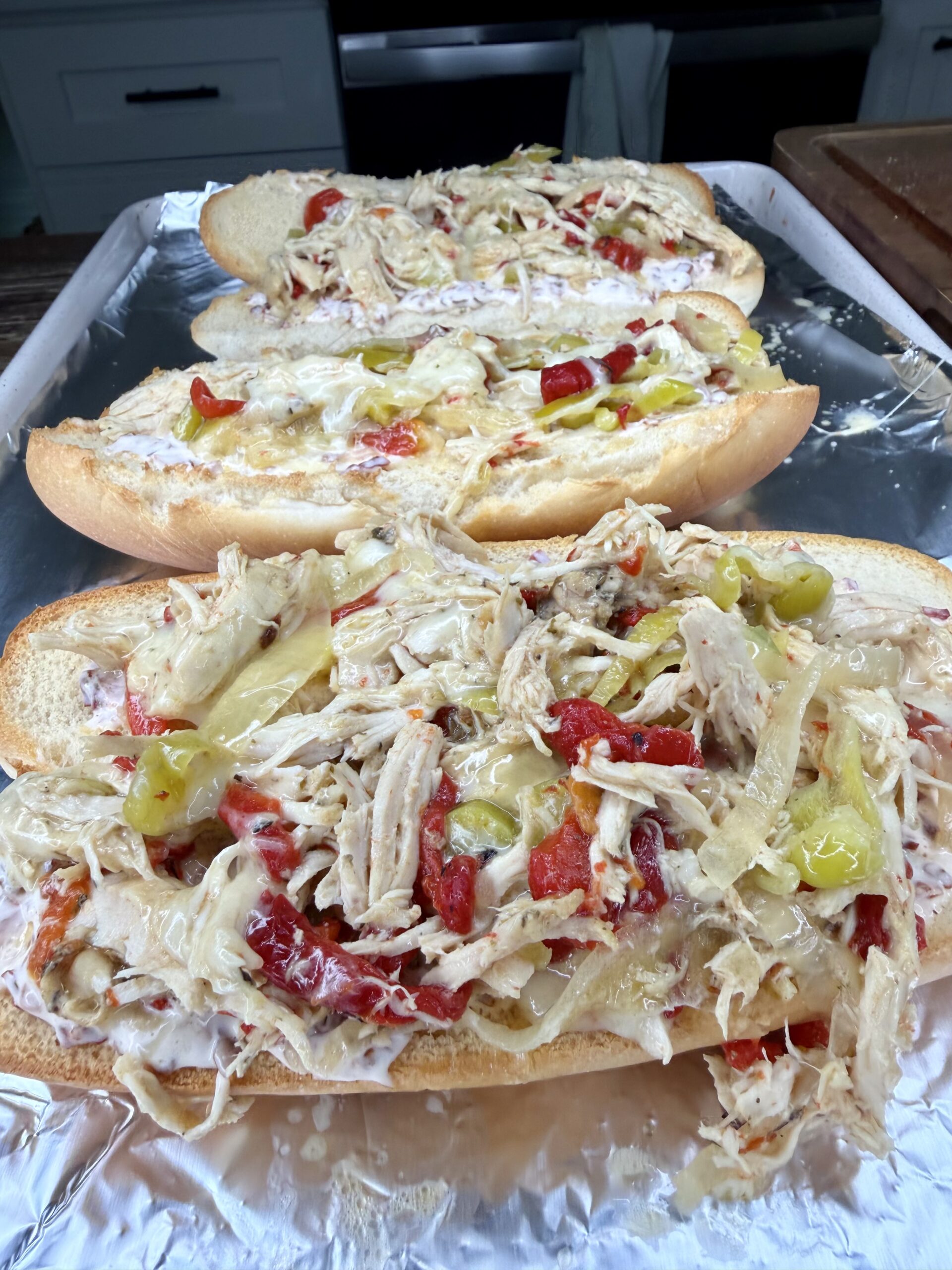 Three open-faced Easy Crockpot Chicken Philly Cheesesteak sandwiches sit on a baking sheet, topped with shredded chicken, sliced peppers, onions, and melted cheese on hoagie rolls, ready to be baked.