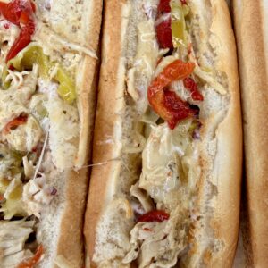 Three toasted hoagie rolls filled with Easy Crockpot Chicken Philly Cheesesteak—shredded chicken, melted cheese, red bell peppers, and banana peppers—are arranged closely together on a foil surface.