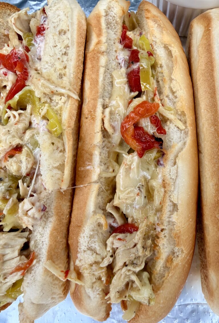 Three toasted hoagie rolls filled with Easy Crockpot Chicken Philly Cheesesteak—shredded chicken, melted cheese, red bell peppers, and banana peppers—are arranged closely together on a foil surface.