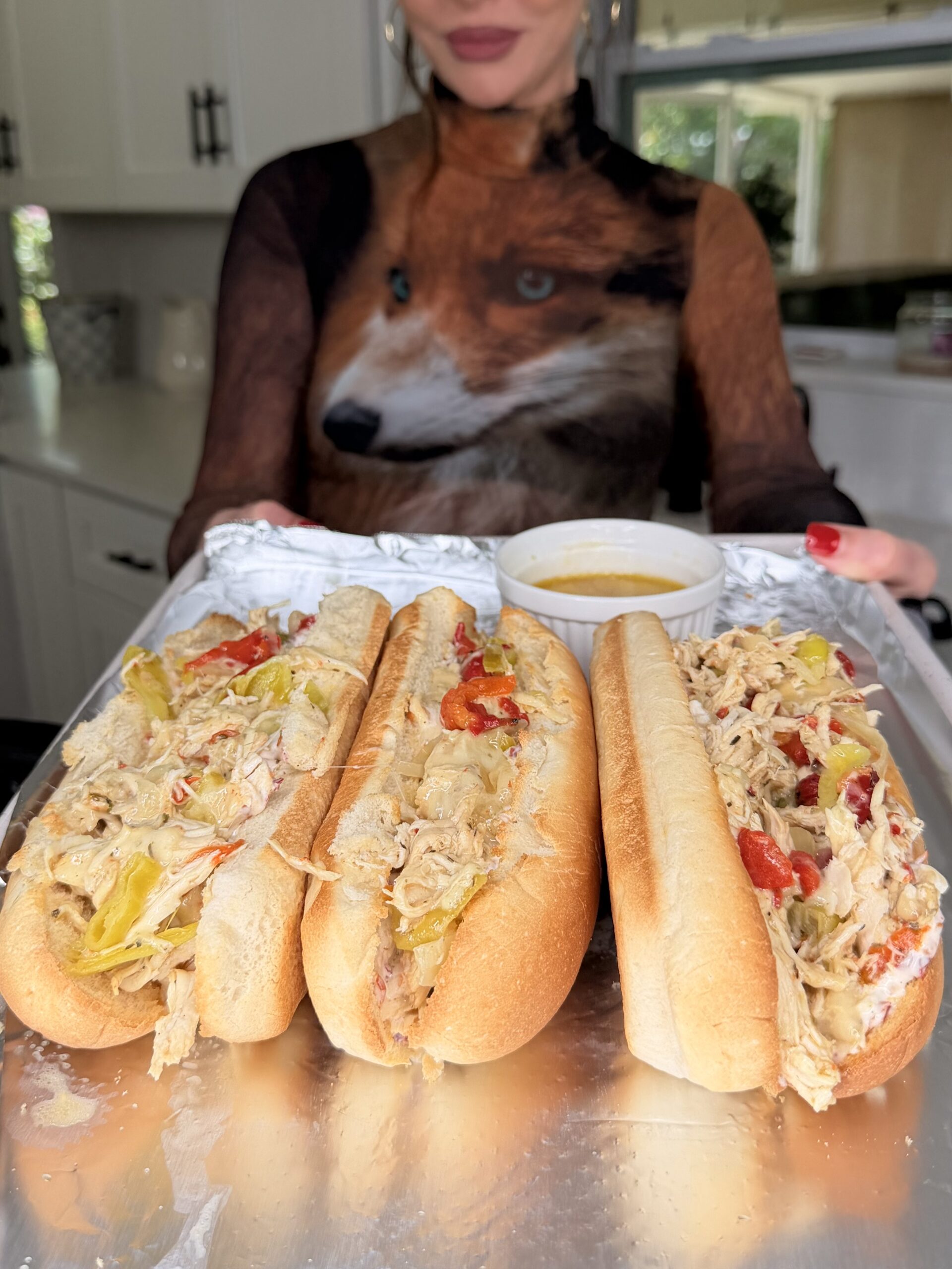 A woman in a shirt with a fox design holds a tray of Easy Crockpot Chicken Philly Cheesesteak hoagie rolls filled with shredded chicken, peppers, and cheese, with a small bowl of dipping sauce in a cozy kitchen setting.