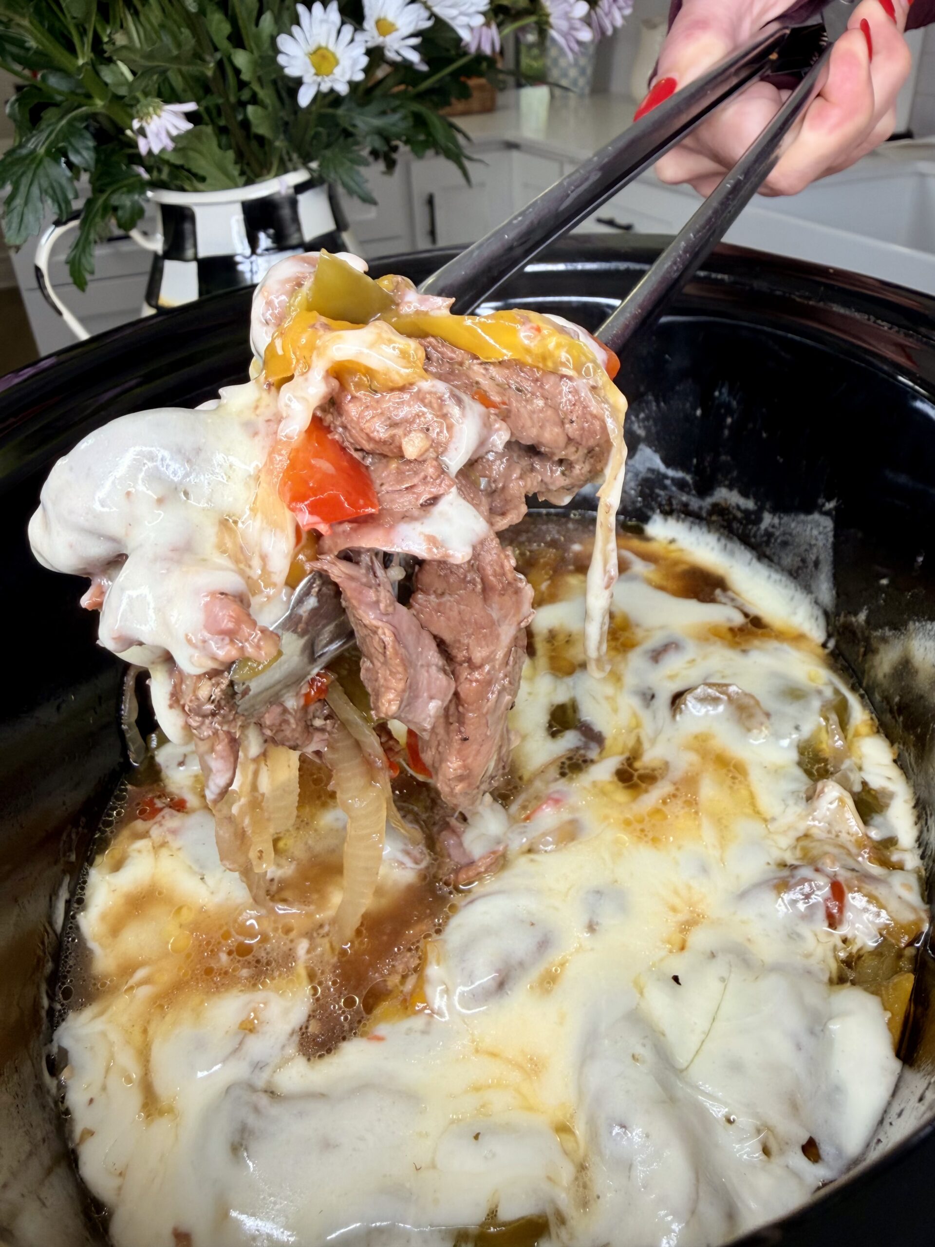 A close-up of chopsticks lifting cheesy Easy Crockpot Philly Cheesesteaks from a slow cooker, with melted cheese, beef, peppers, and onions. Flowers and part of a kitchen are visible in the background.