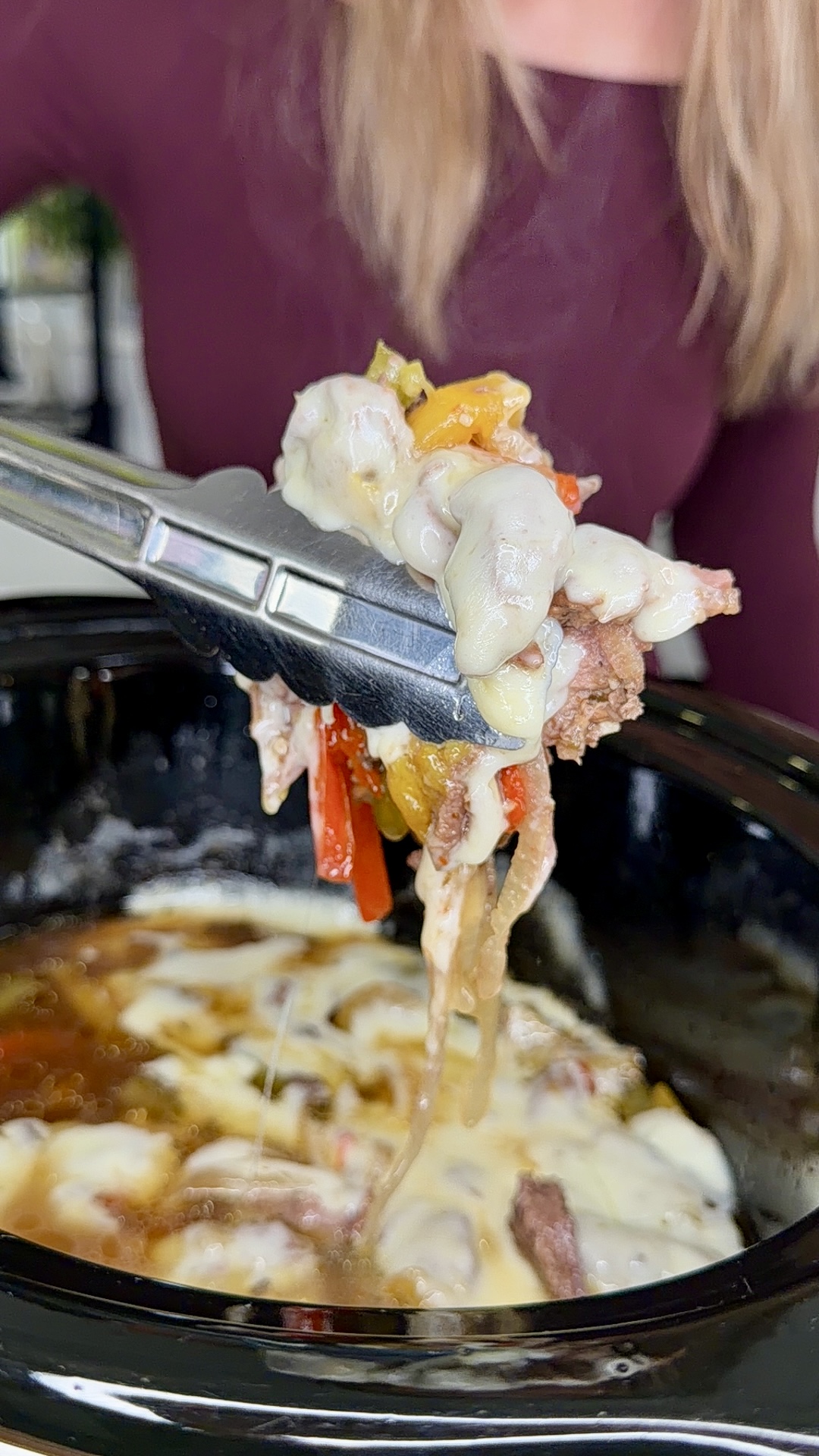 A person uses metal tongs to lift a portion of melted cheese, sliced beef, and peppers from a slow cooker filled with Easy Crockpot Philly Cheesesteaks—a cheesy, savory dish.