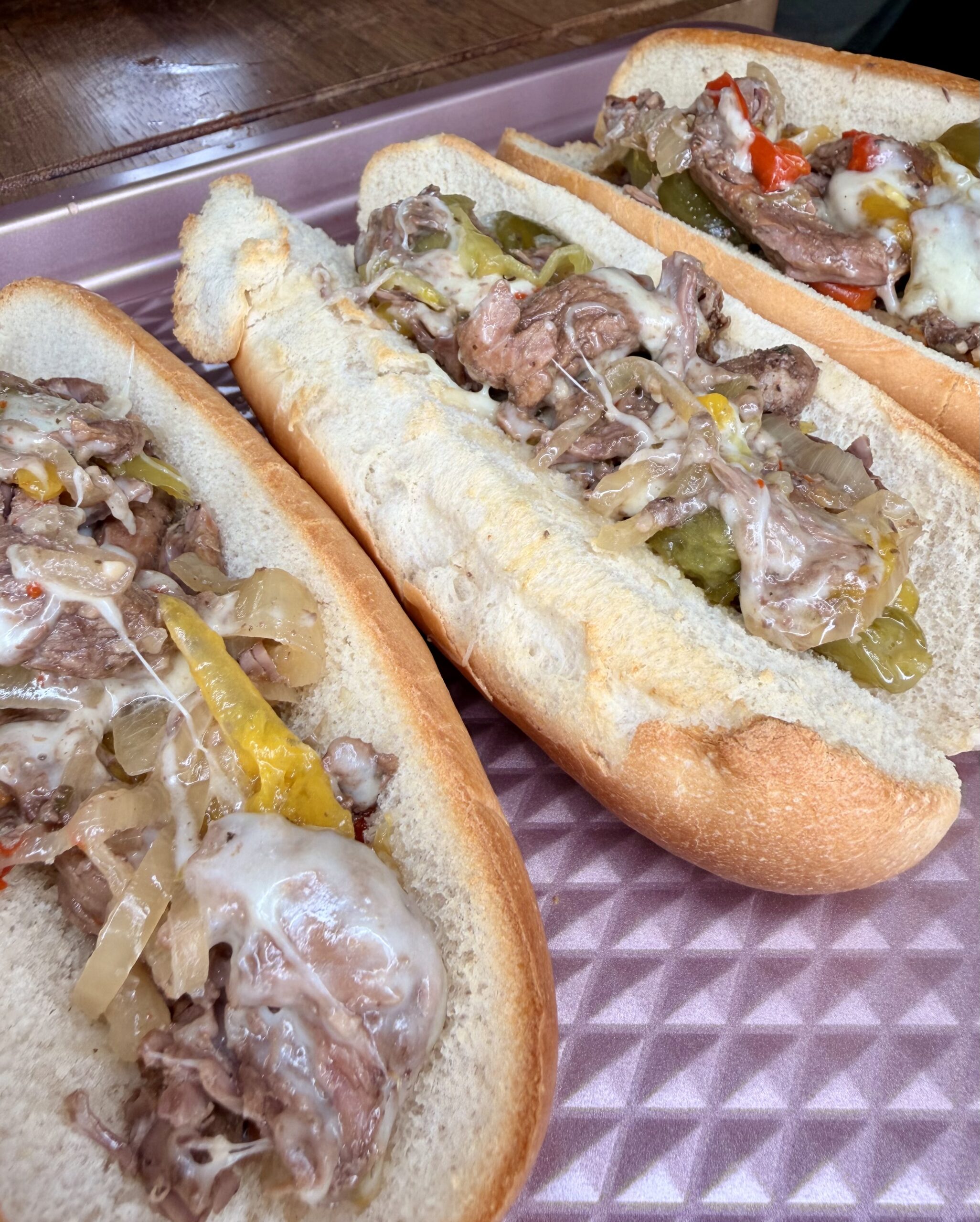 Three Easy Crockpot Philly Cheesesteaks on hoagie rolls, filled with sliced beef, melted cheese, sautéed onions, and peppers, served on a pink tray.