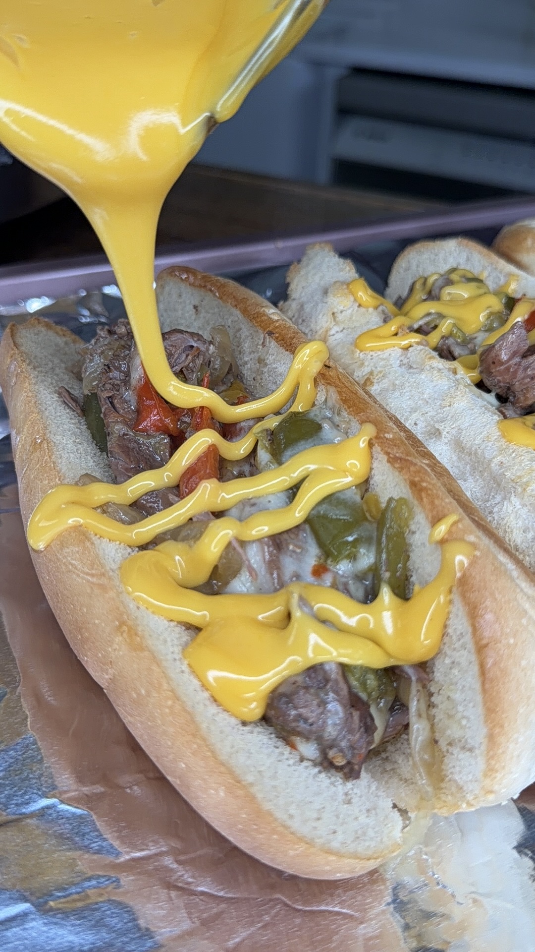 Close-up of melted cheese poured onto a sandwich roll filled with sliced steak, onions, and green peppers—just like Easy Crockpot Philly Cheesesteaks. More cheese is drizzled on top as the hearty sandwich rests on foil.