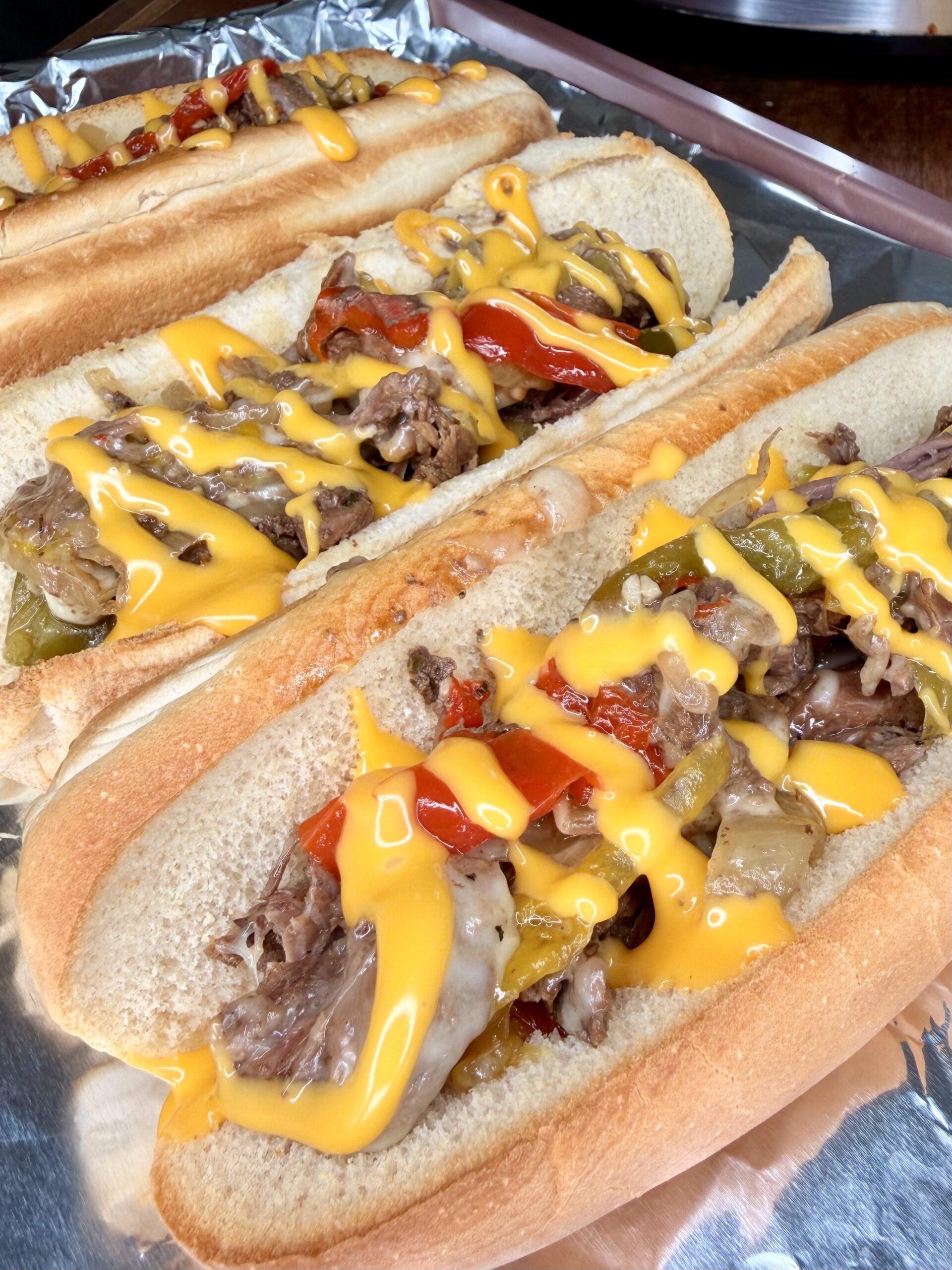 Three hoagie rolls filled with sliced beef, sautéed red and green bell peppers, onions, and topped with melted yellow cheese sauce, arranged on a sheet of aluminum foil—just like classic Easy Crockpot Philly Cheesesteaks.