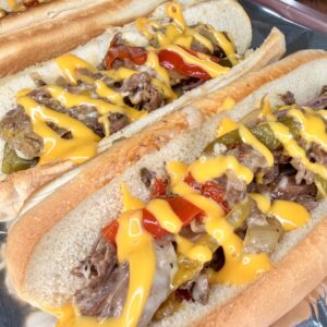 Three Easy Crockpot Philly Cheesesteaks on hoagie rolls, filled with sliced beef, sautéed onions, red and green bell peppers, and topped with melted yellow cheese sauce, all served on a sheet of aluminum foil.