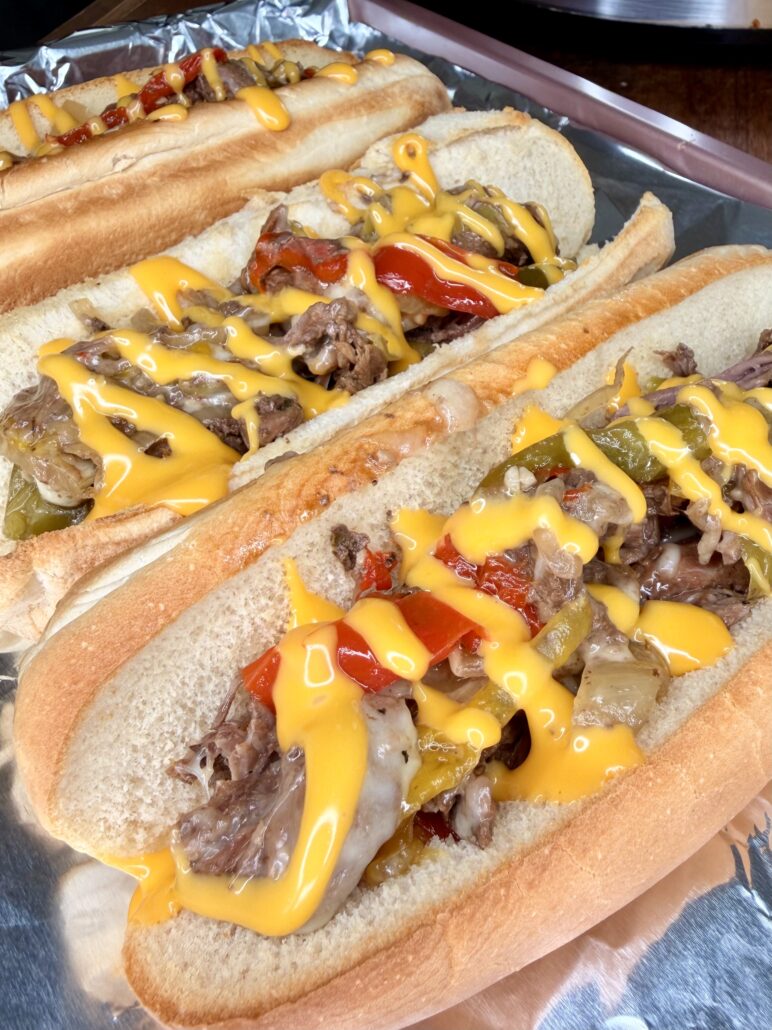 Three Easy Crockpot Philly Cheesesteaks on hoagie rolls, filled with sliced beef, sautéed onions, red and green bell peppers, and topped with melted yellow cheese sauce, all served on a sheet of aluminum foil.