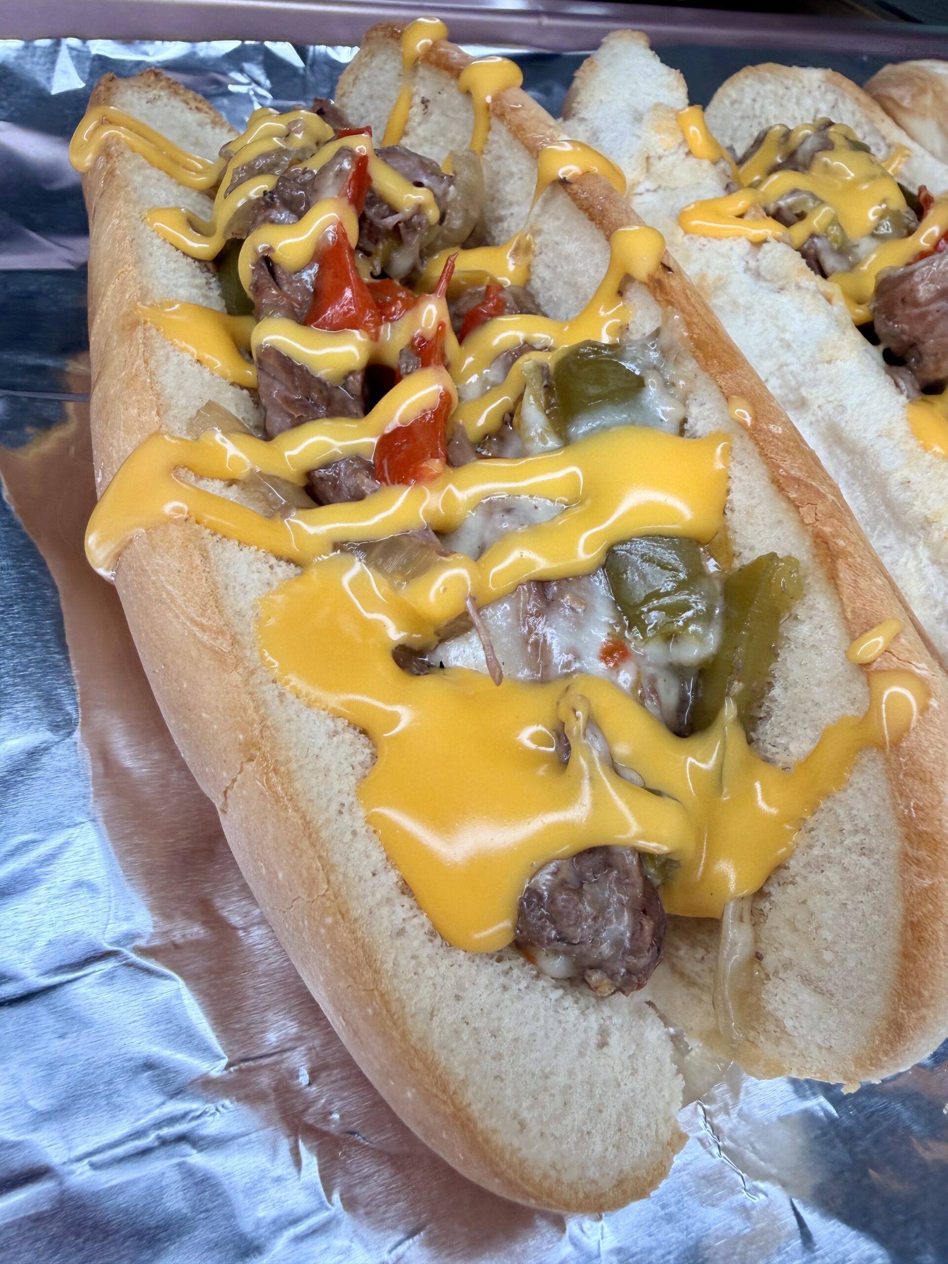 A close-up of an Easy Crockpot Philly Cheesesteaks sandwich with grilled sausage, sautéed onions, red and green bell peppers, all topped with melted cheddar cheese sauce, served in a hoagie roll on aluminum foil.