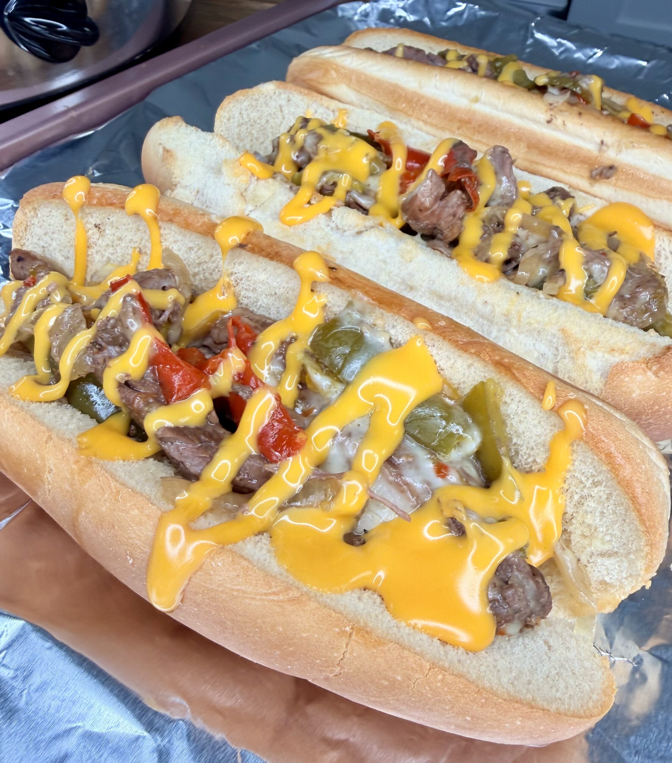 Three hoagie rolls filled with sliced steak, sautéed onions, red and green bell peppers, topped with melted yellow cheese sauce—these Easy Crockpot Philly Cheesesteaks are arranged perfectly on foil for a mouthwatering meal.