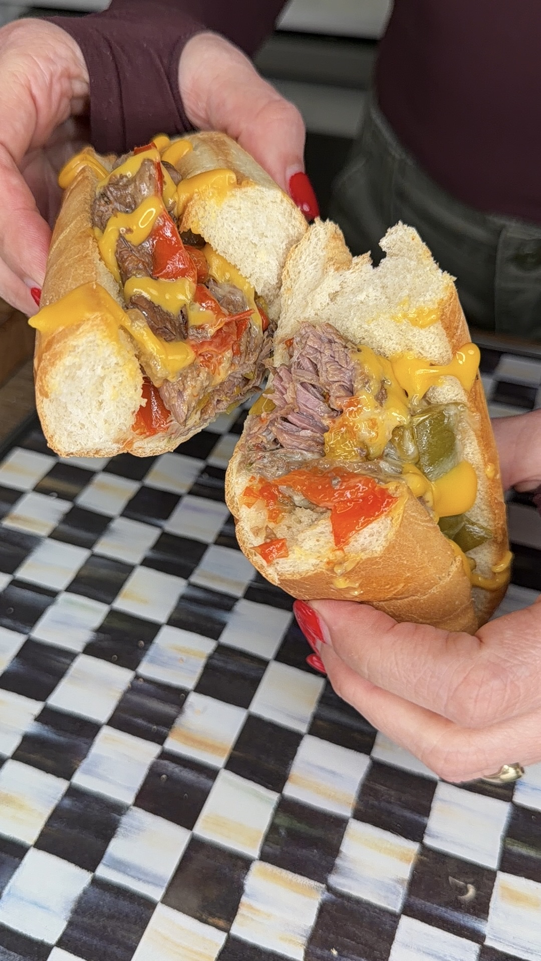 Two hands hold a split sandwich filled with shredded beef, melted cheese, and sautéed red and green peppers—an Easy Crockpot Philly Cheesesteaks creation—over a checkered black, white, and gray tile surface.