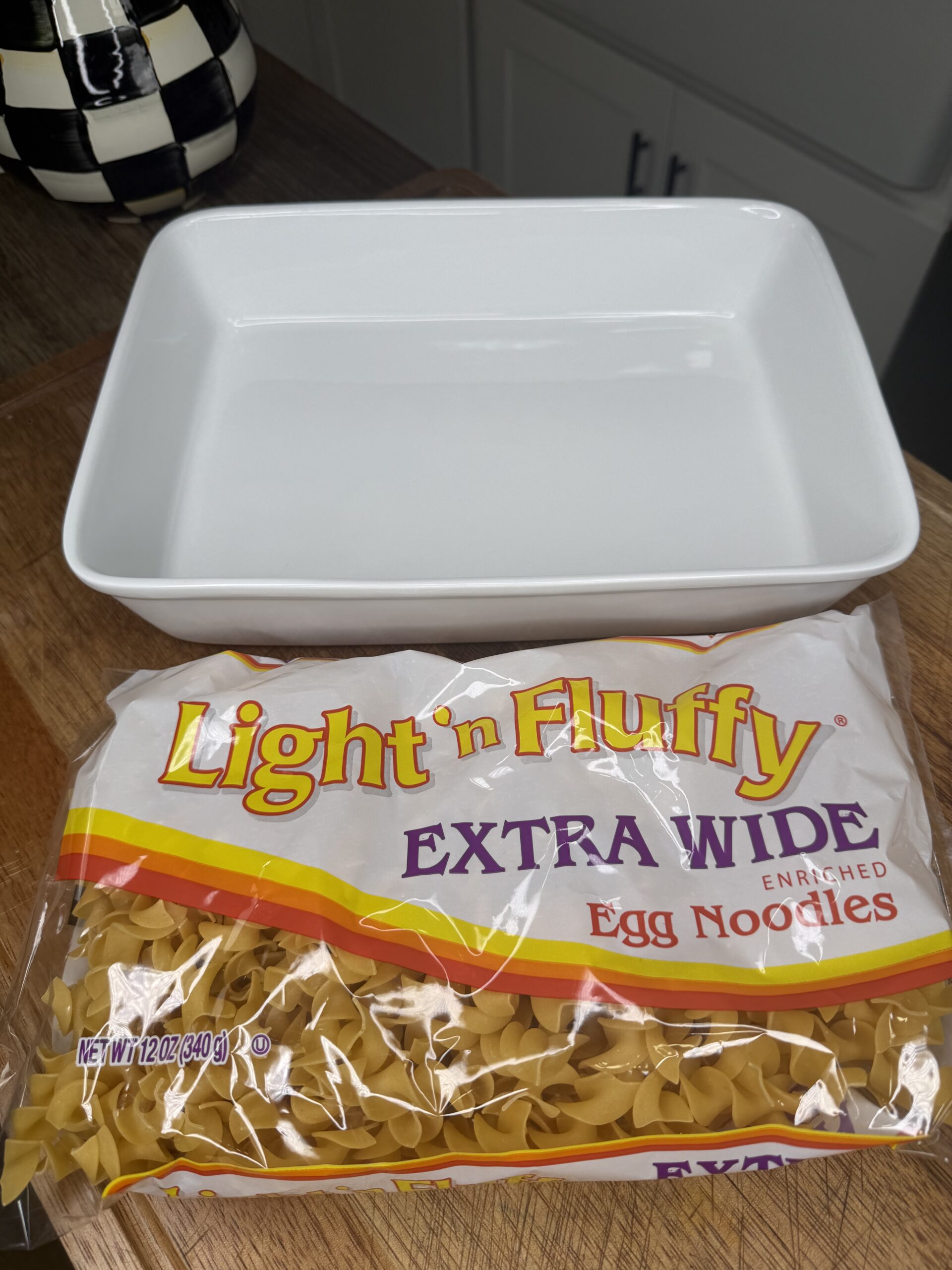 A white rectangular baking dish sits on a wooden surface behind an unopened bag of Light n Fluffy Extra Wide Egg Noodles.