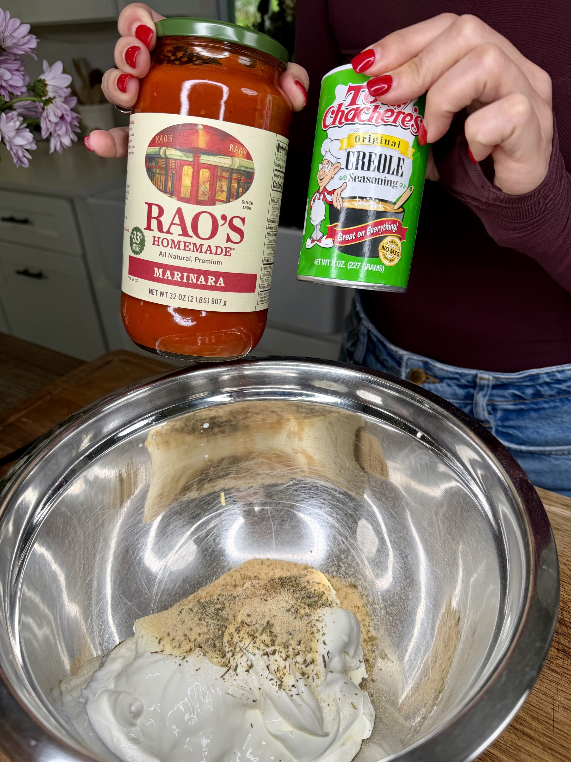 A person holds a jar of Rao’s Homemade Marinara sauce and a can of Tony Chachere’s Creole Seasoning above a metal bowl containing white sauce and various spices.