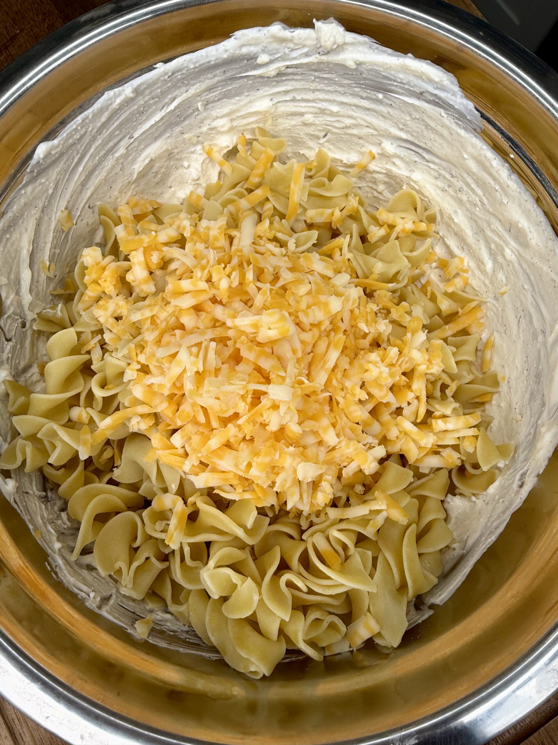 A large metal bowl filled with cooked egg noodles, shredded yellow and white cheese, and a creamy sauce coating the sides of the bowl.