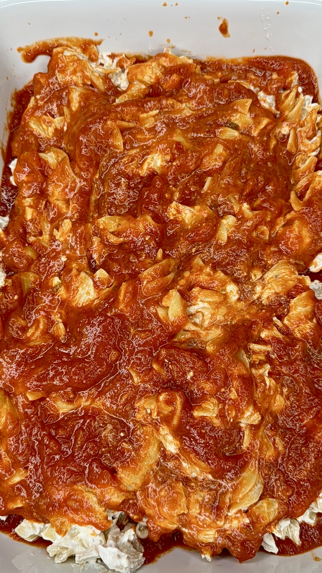 A close-up of a dish with a layer of bright red tomato sauce spread unevenly over chopped vegetables or pasta, possibly part of a homemade lasagna or casserole in a rectangular pan.