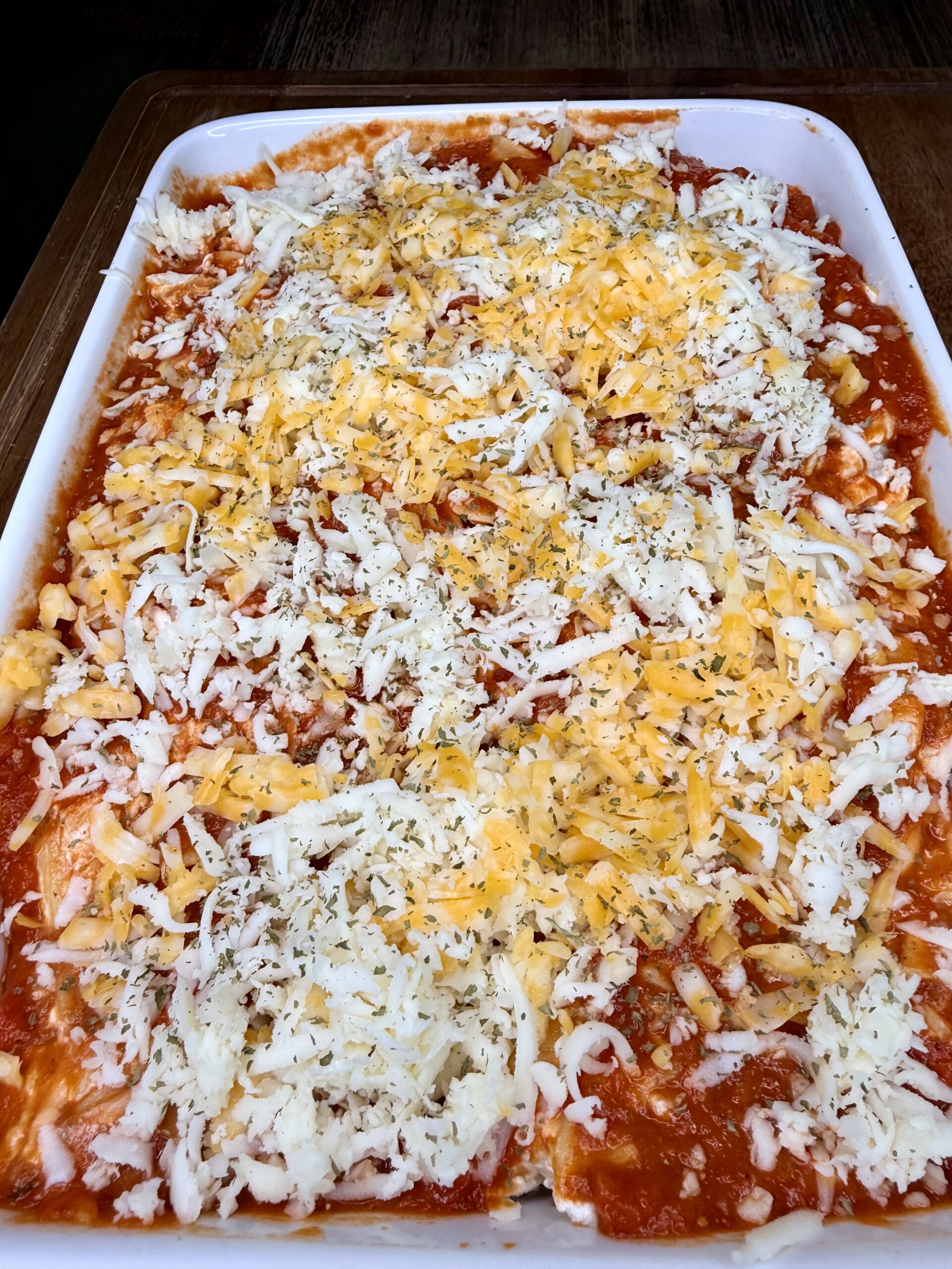 A large rectangular baking dish filled with lasagna topped with red tomato sauce, shredded mozzarella and cheddar cheese, and sprinkled herbs, ready to be baked.