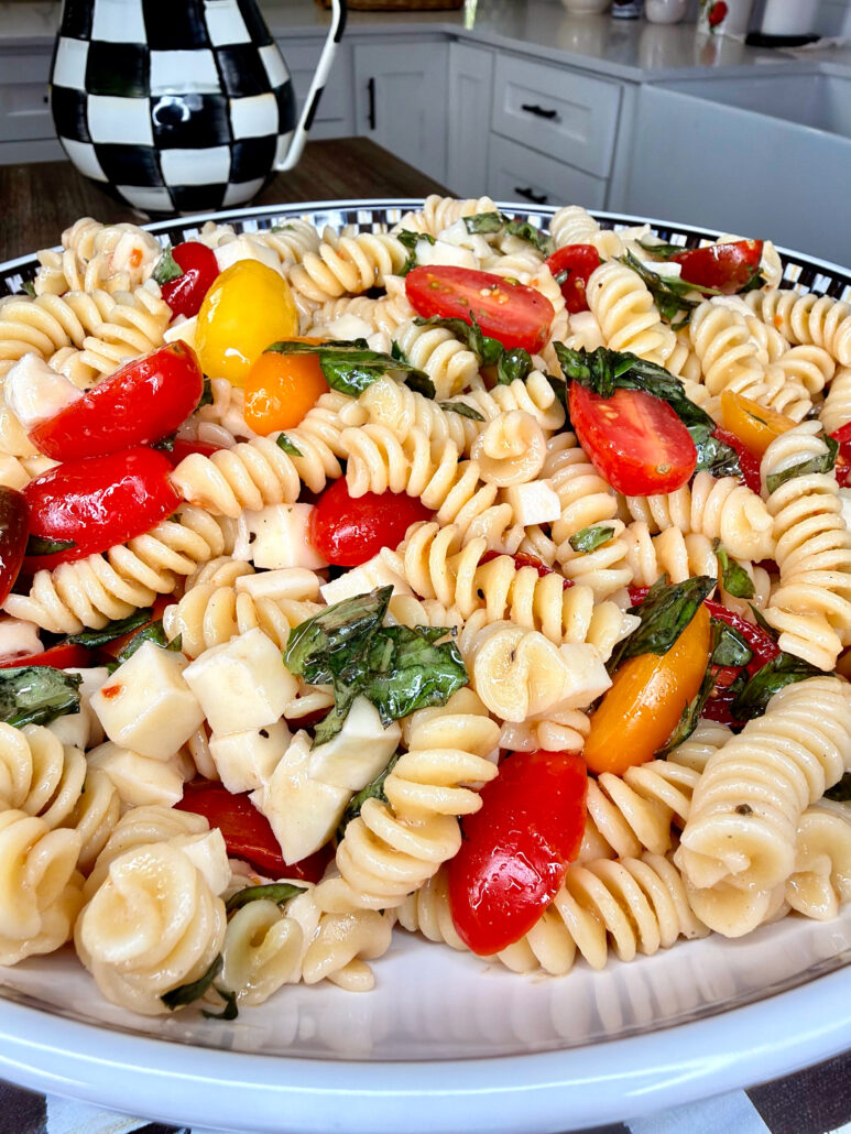 A close-up of an Easy Italian Pasta Salad featuring rotini, cherry tomatoes, mozzarella cubes, and fresh basil on a kitchen counter. The background showcases a checkered teapot and a farmhouse-style sink.