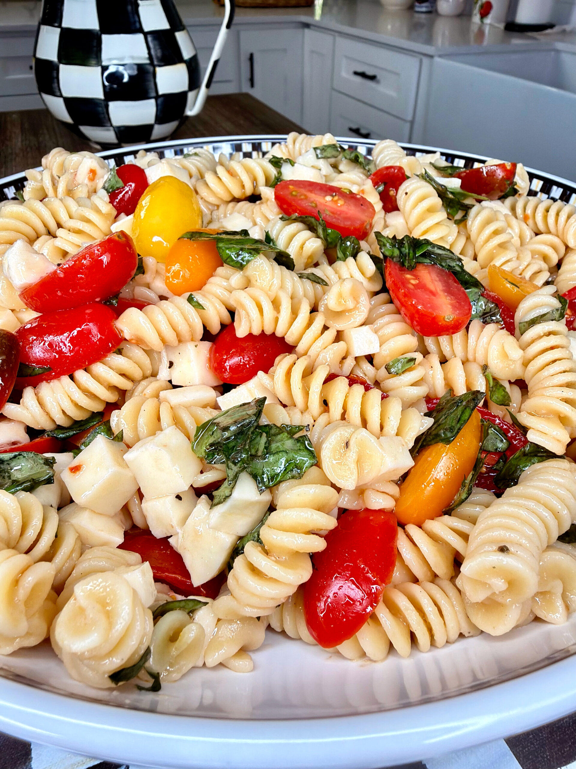A close-up of an Easy Italian Pasta Salad featuring rotini, cherry tomatoes, mozzarella cubes, and fresh basil on a kitchen counter. The background showcases a checkered teapot and a farmhouse-style sink.