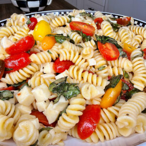 A close-up of an Easy Italian Pasta Salad with rotini, cherry tomatoes, basil leaves, and mozzarella chunks served in a decorative bowl in a kitchen setting.