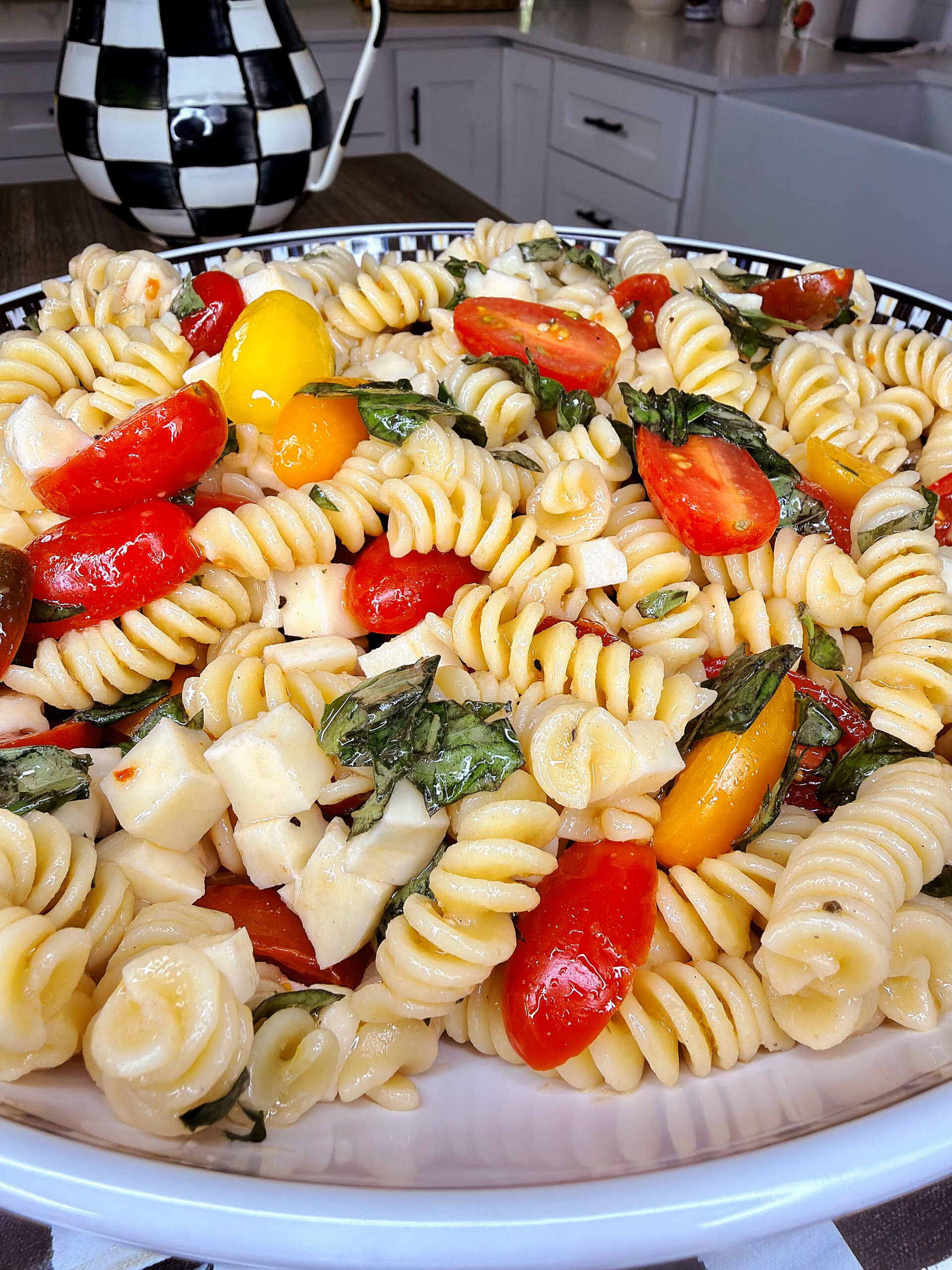A bowl of Easy Italian Pasta Salad with rotini, halved cherry tomatoes, fresh basil leaves, and cubes of mozzarella cheese sits on a kitchen table.