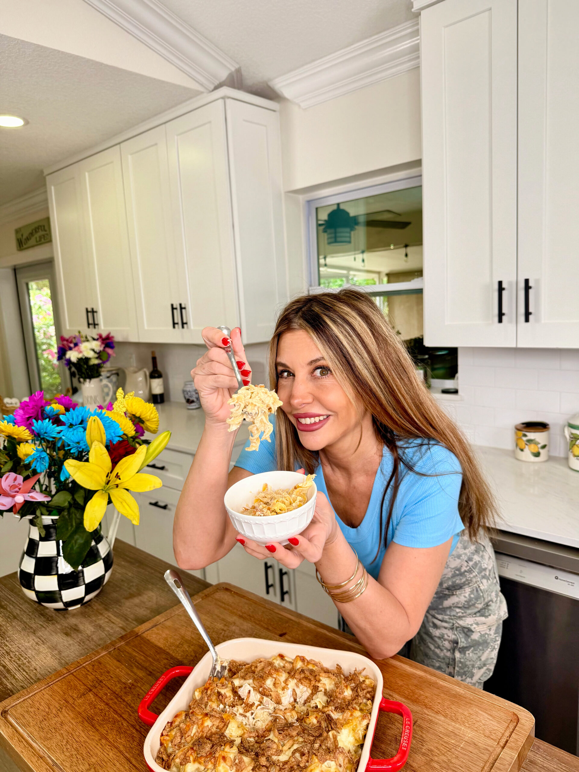 A smiling woman in a blue shirt holds up a bowl of food with a fork in a bright kitchen. A casserole dish sits on the counter beside her, and a colorful bouquet of flowers is in the background.
