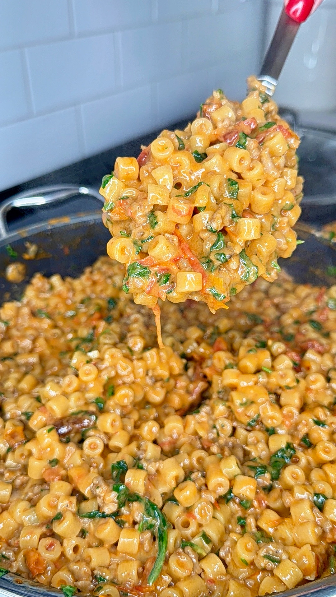 A close-up of creamy ditalini pasta mixed with ground meat, tomatoes, and spinach, being scooped from a pan with a spoon in a kitchen setting.