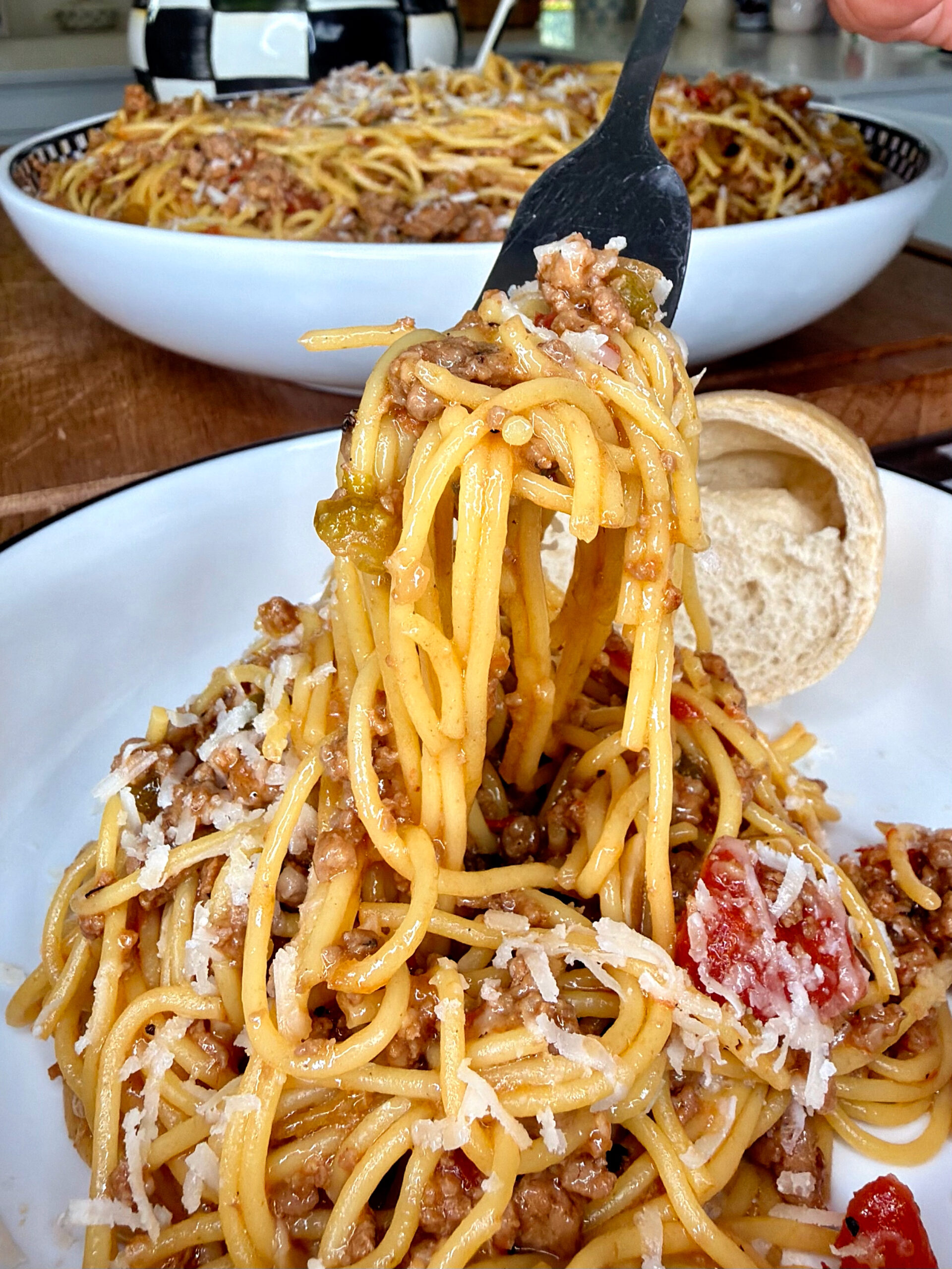 A close-up of a fork lifting a serving of spaghetti with meat sauce and grated cheese from a white plate, with a bread roll on the side and a large bowl of pasta in the background.