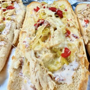 A toasted hoagie roll topped with Easy Crockpot Chicken Philly Cheesesteak filling—shredded chicken, melted cheese, sliced pickles, chopped red peppers, and a creamy white sauce—all placed on aluminum foil.