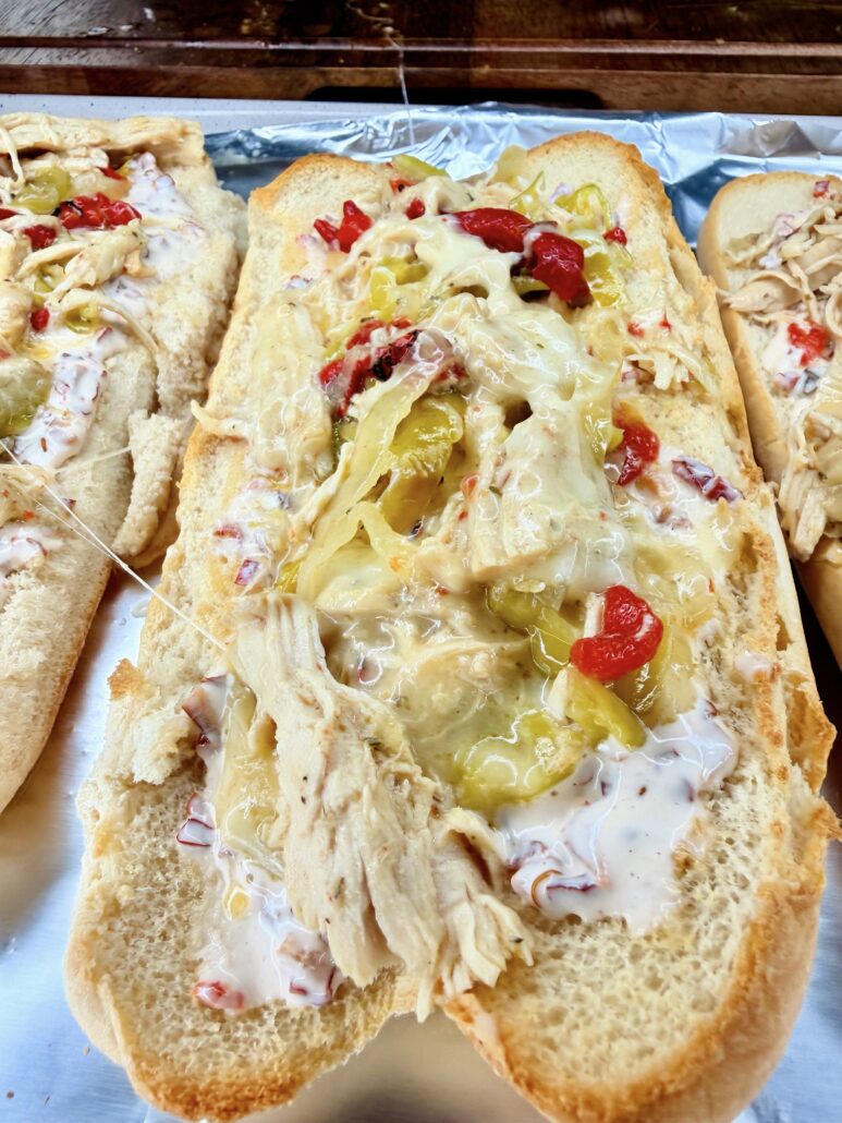 A toasted hoagie roll topped with Easy Crockpot Chicken Philly Cheesesteak filling—shredded chicken, melted cheese, sliced pickles, chopped red peppers, and a creamy white sauce—all placed on aluminum foil.