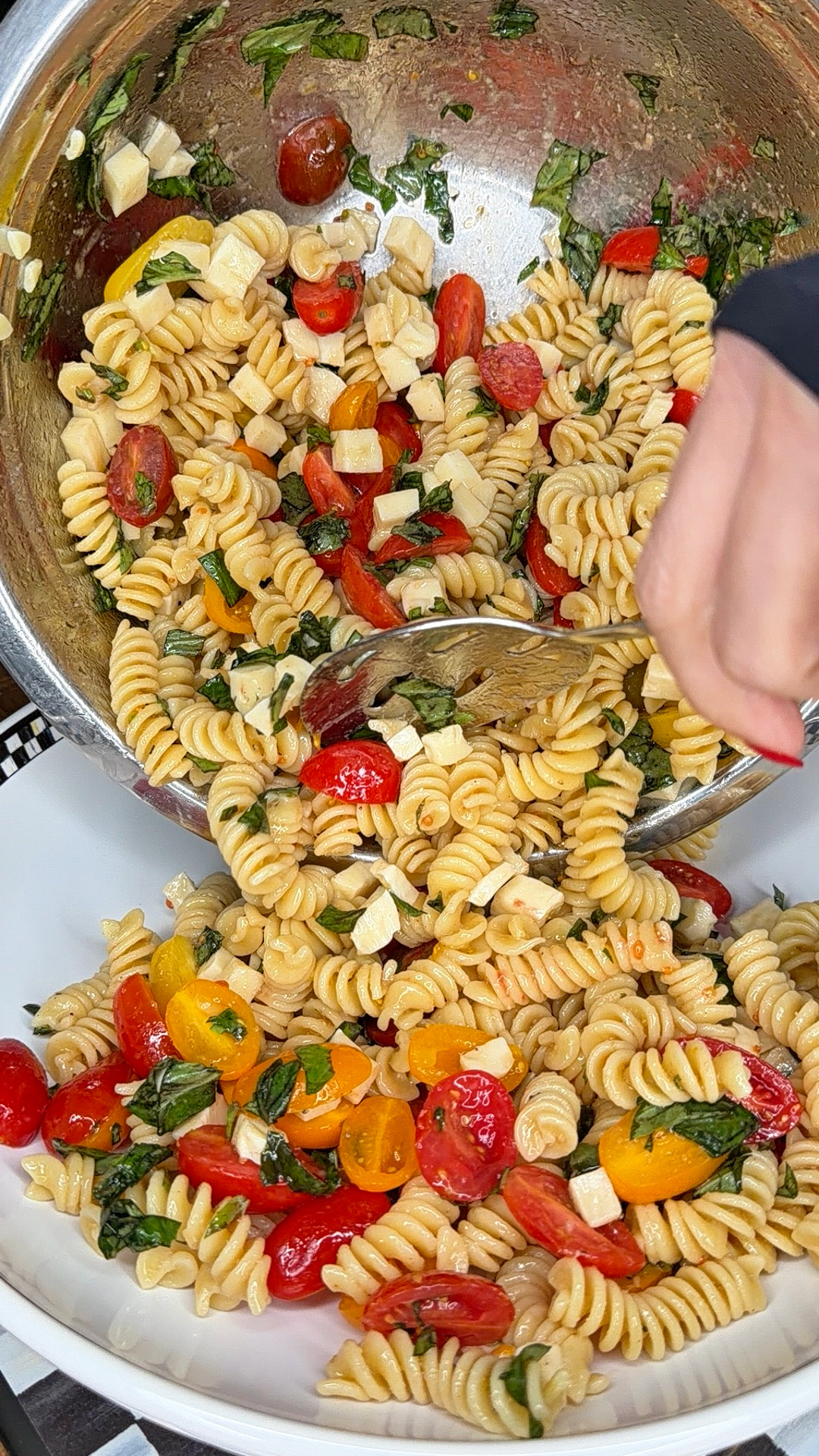 A hand serves Easy Italian Pasta Salad—rotini with cherry tomatoes, mozzarella cubes, and fresh basil—from a large metal mixing bowl into a white dish.