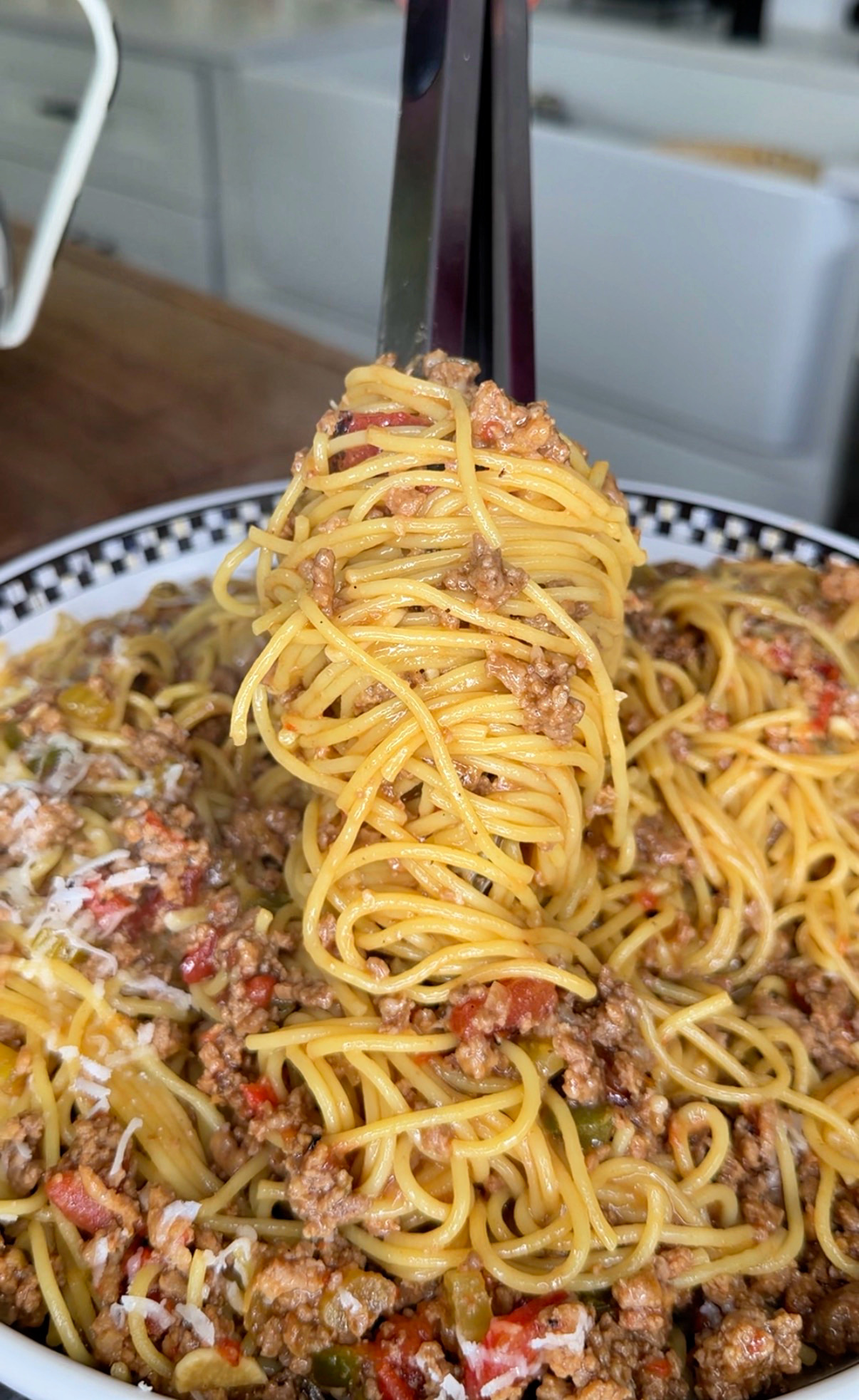 A close-up of spaghetti with meat sauce being twirled by metal tongs above a bowl, showing ground meat, tomato chunks, and grated cheese mixed into the pasta.