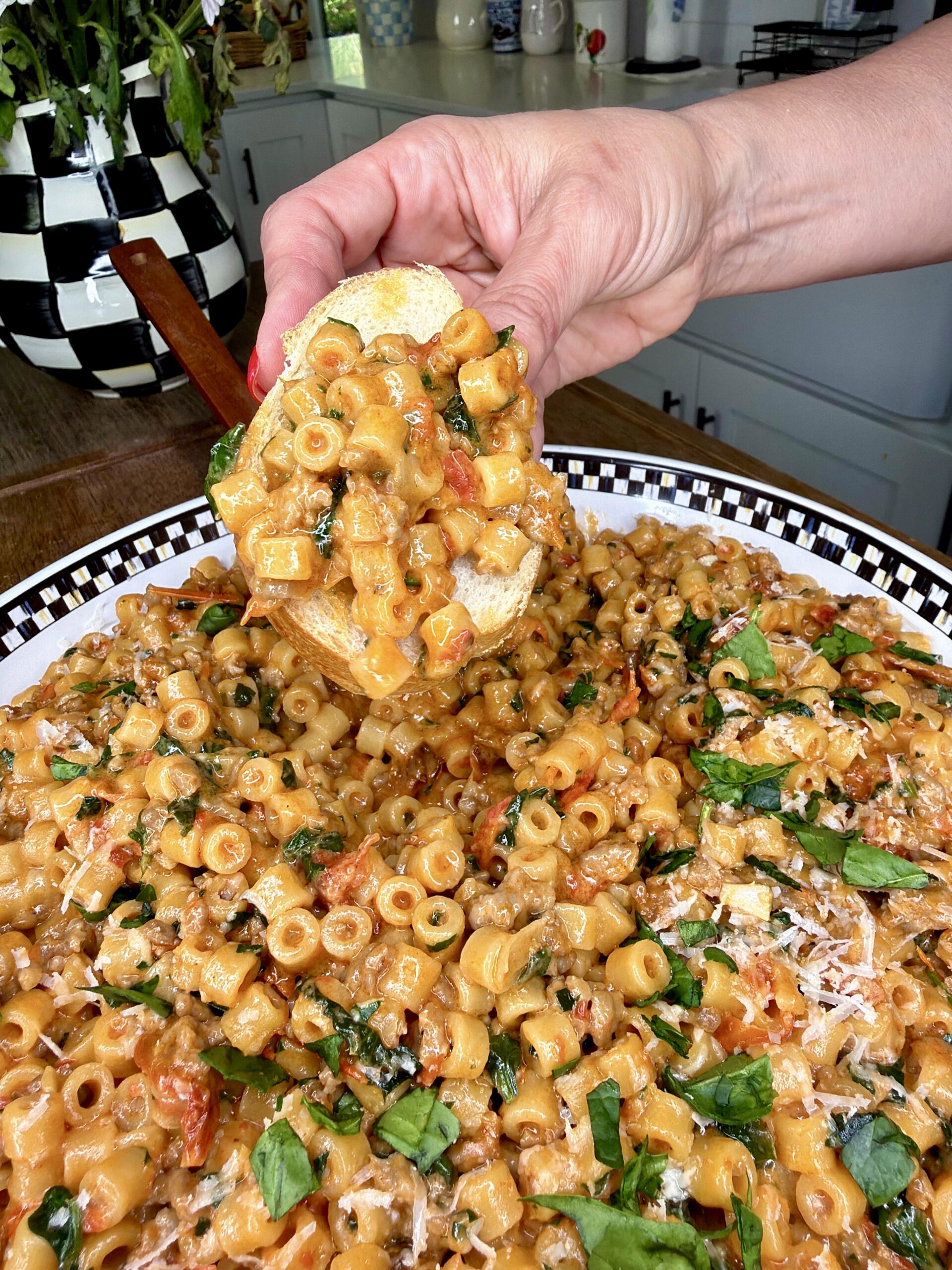 A hand holds a slice of bread topped with Sicilian One-Pot Creamy Sausage Pasta, garnished with fresh basil, above a large checkered platter filled with more of this delicious pasta in a bright kitchen setting.